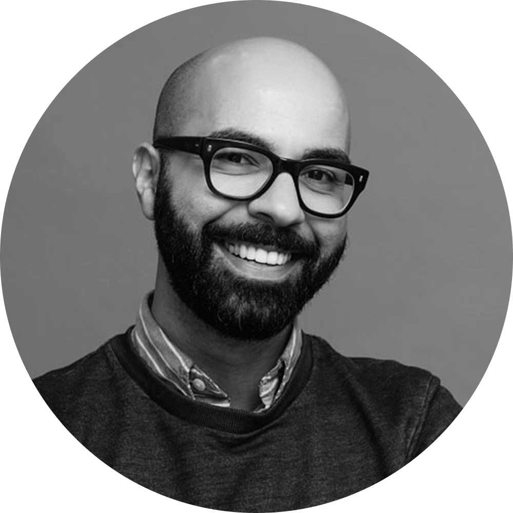 Black and white photo of a smiling man with a beard, glasses, wearing a sweater over a collared shirt.