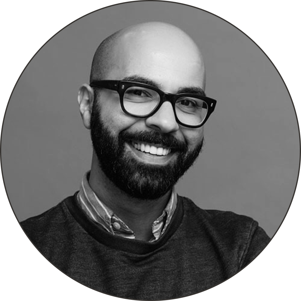 A black and white portrait of a smiling Amer with glasses, a beard, and a striped collared shirt, wearing a sweater.