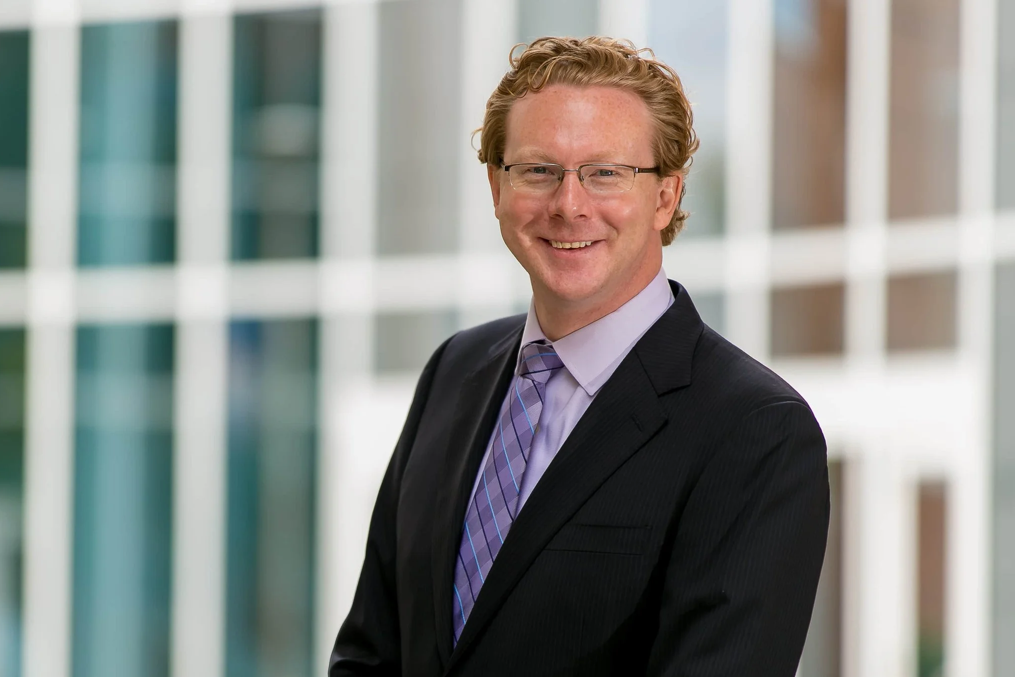 A smiling man with curly red hair and glasses dressed in a dark suit, light purple shirt, and purple tie, standing in front of a modern office building with glass windows.