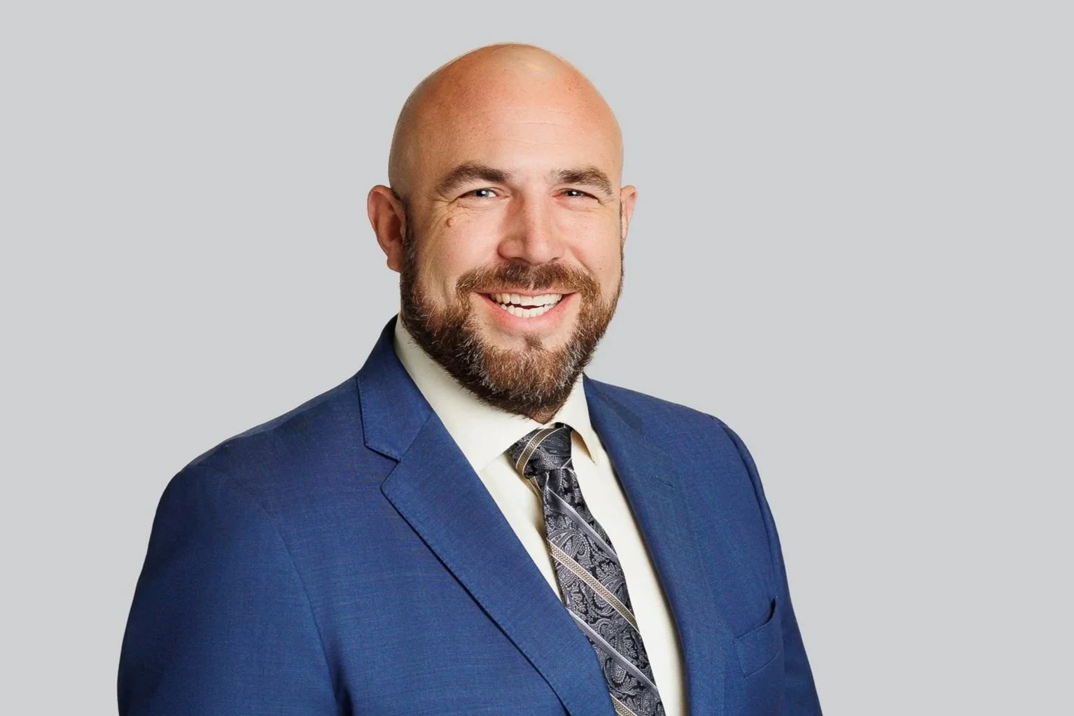 Professional headshot of smiling man with beard in a blue suit and patterned tie against a light gray background