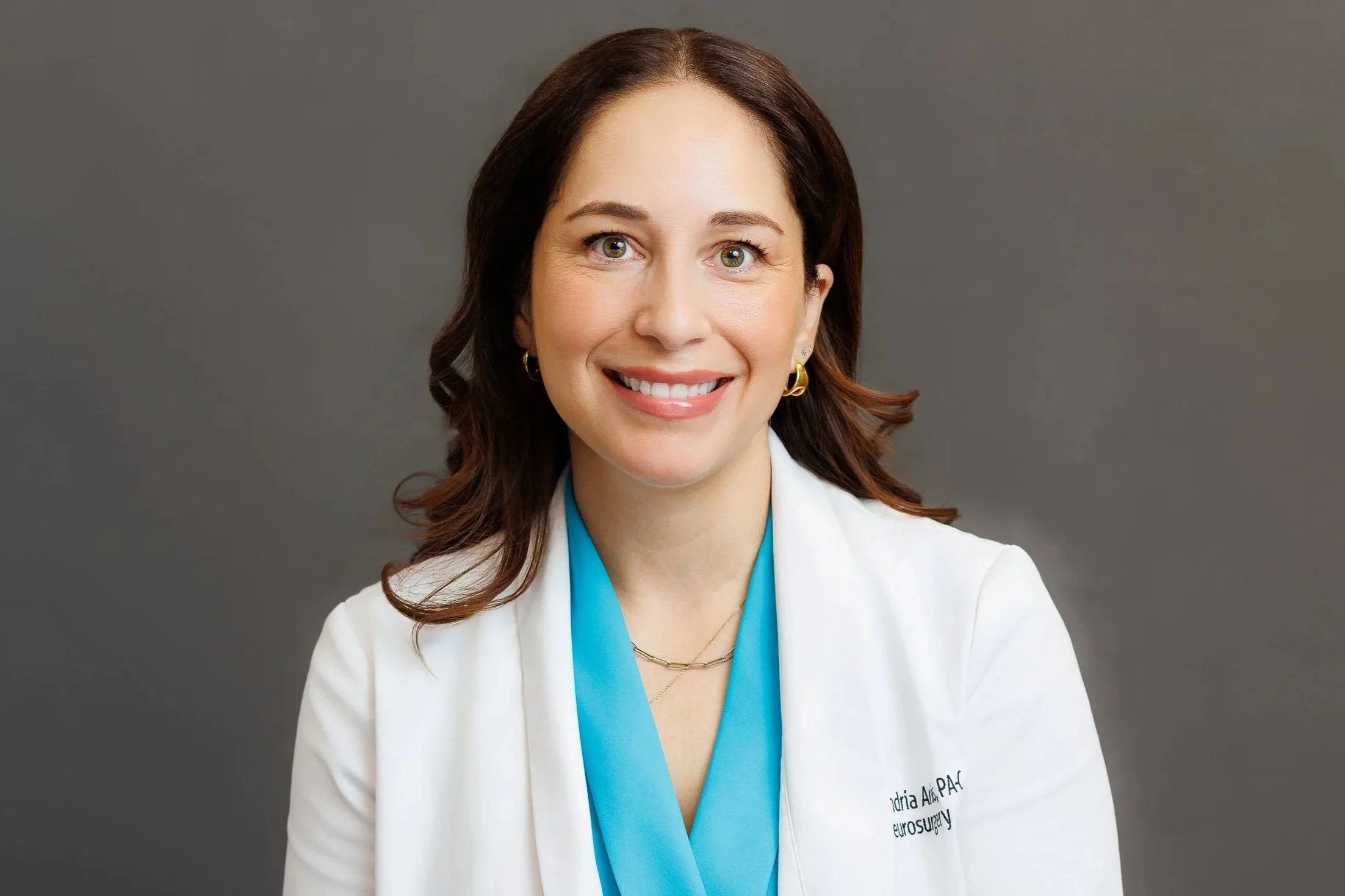 A woman with brown hair and green eyes smiling, dressed in a white medical coat with a blue scrubs underneath, against a plain gray background.