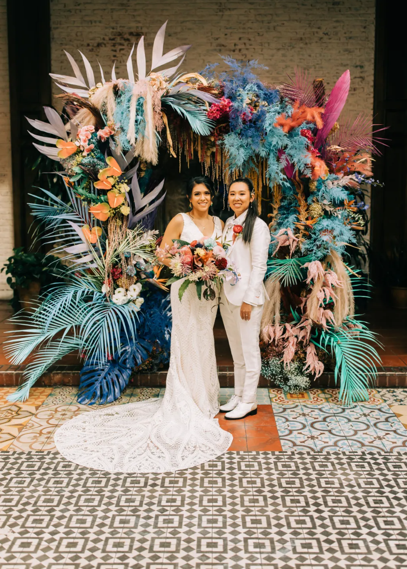 Two women standing in front of a colorful floral arch at a wedding or event. One woman is wearing a white wedding gown and holding a bouquet, while the other is dressed in a white suit. The background features a brick wall and patterned floor tiles.