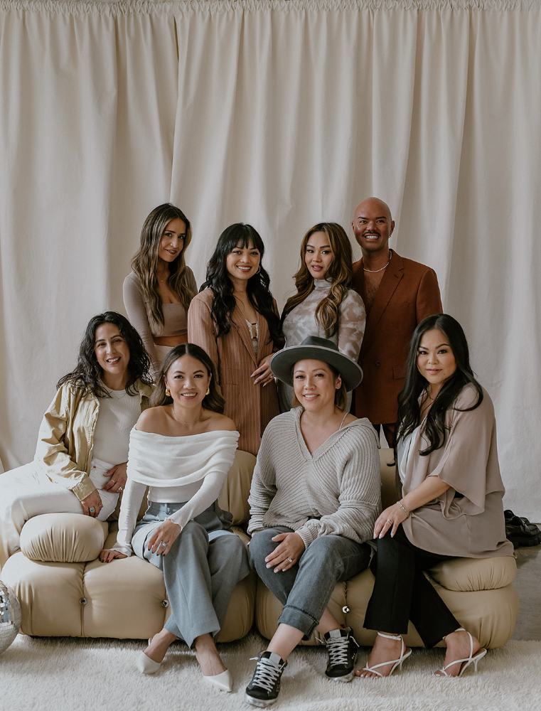 Ten diverse women posing together in a casual indoor setting, some sitting on a beige sofa and others standing behind, against a cream-colored curtain backdrop.