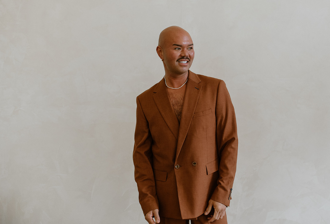 A bald man with a mustache and earring wearing a brown suit, necklace, and no shirt underneath, standing against a plain light-colored wall, smiling and looking to the side.