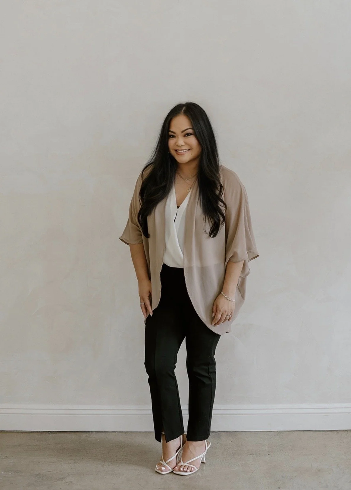 Smiling woman with long black hair wearing a beige kimono over a white blouse and black pants standing against a plain off-white wall.