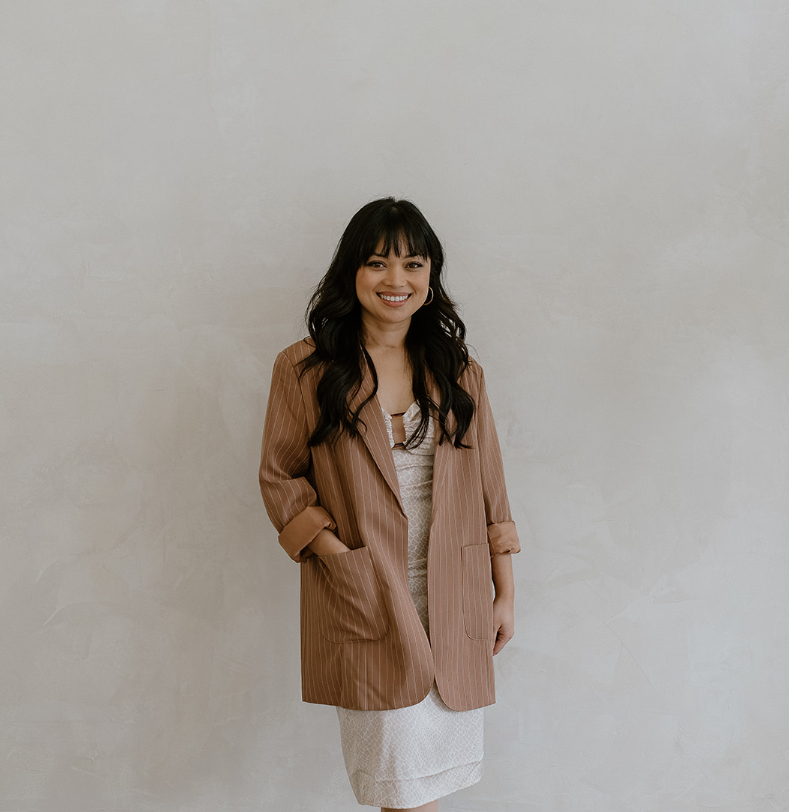 A woman with long dark hair and bangs standing against a plain light-colored wall, wearing a tan striped blazer over a beige top.