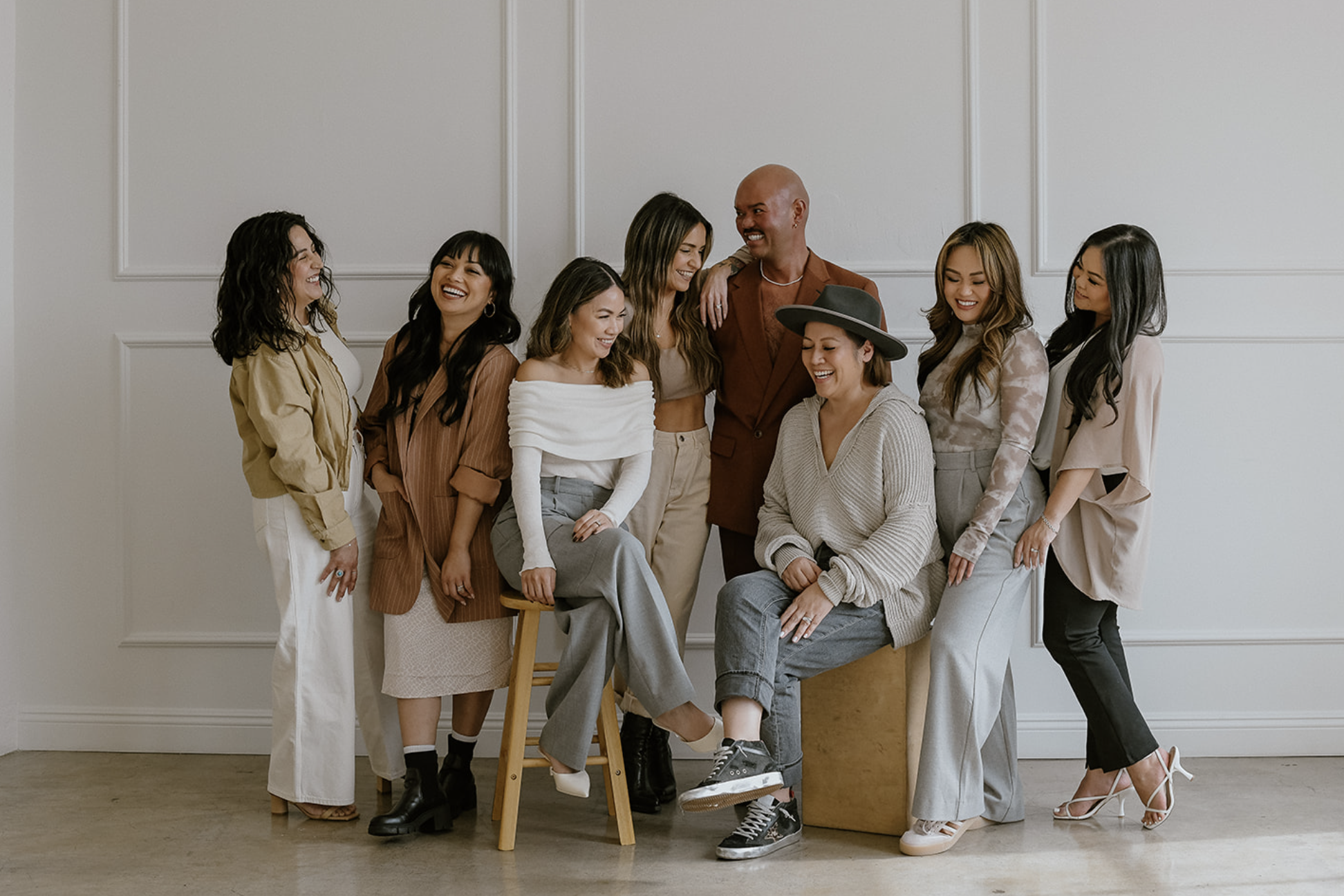 Group of nine diverse women and one man laughing and smiling together indoors against a plain white wall.