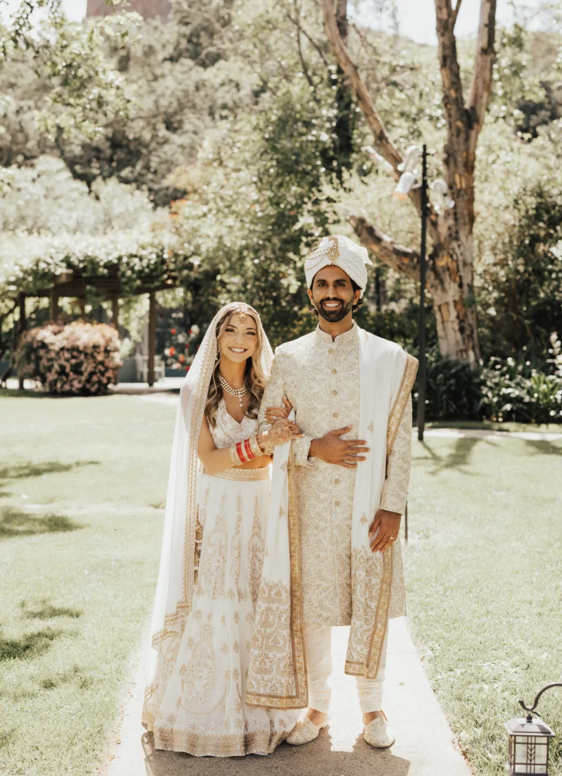 A newlywed Indian couple dressed in traditional wedding attire, standing outdoors on a sunny day with trees and greenery in the background, smiling happily.