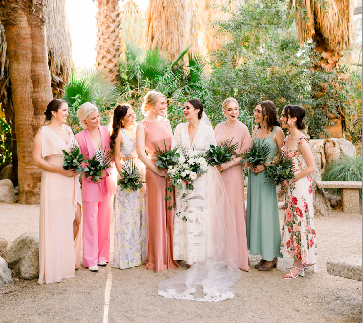 Group of women in colorful dresses at a wedding ceremony, standing outdoors near palm trees and rocks, holding bouquets, with the bride in a white dress and veil, smiling and talking among friends.