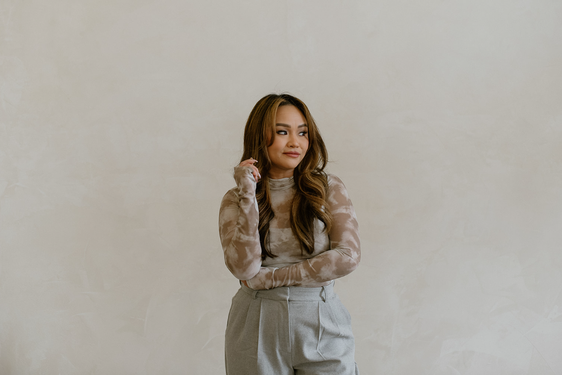 A woman with long wavy hair standing against a plain neutral wall, wearing a patterned long-sleeve top and high-waisted gray trousers.