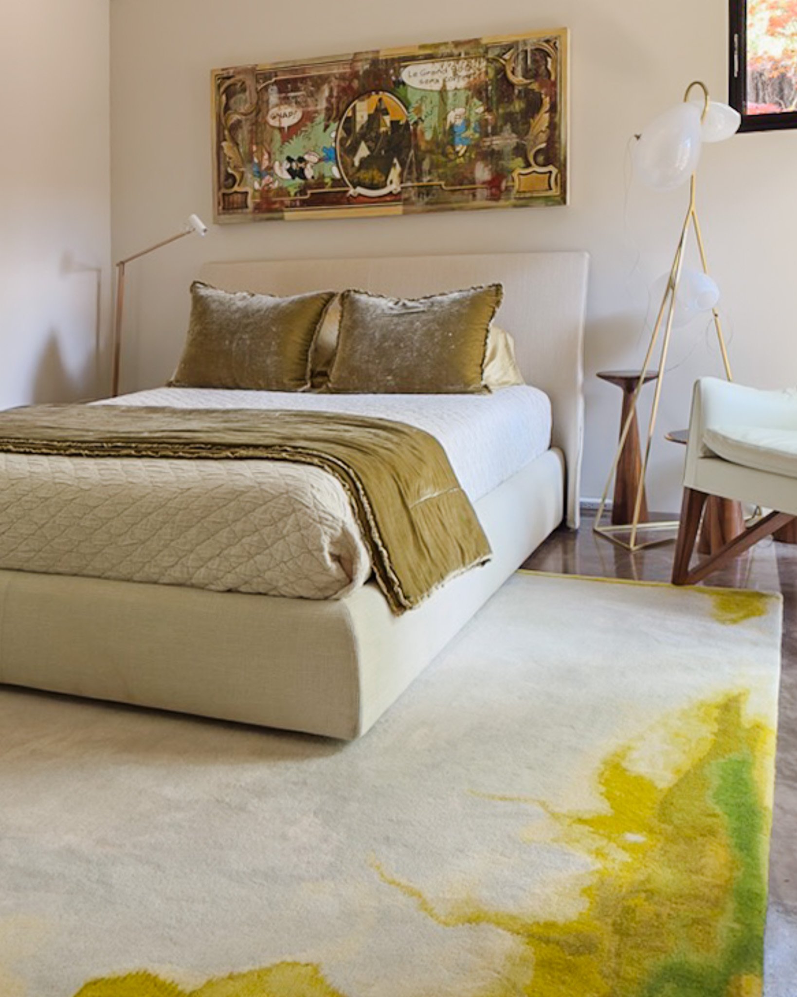 Bedroom with gold and beige bedding, a white headboard, and colorful abstract art, with views of autumn foliage.
