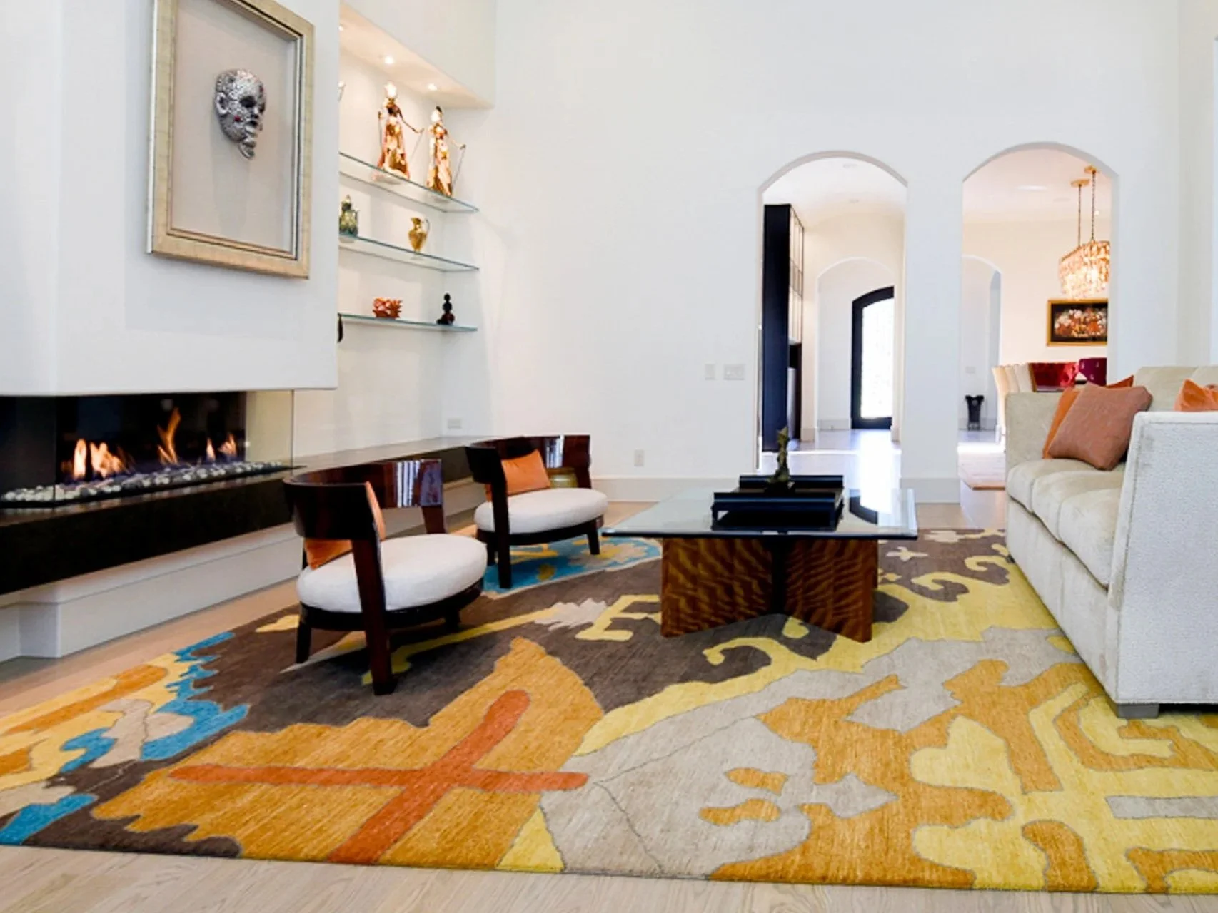 Living room with a patterned multicolored rug, a white sofa with orange pillows, a glass coffee table, and two chairs with dark wood frames and white cushions. A modern fireplace and decorative shelves with sculptures and figurines are on the wall. Arched doorways lead to other rooms, and a chandelier is visible in the background.