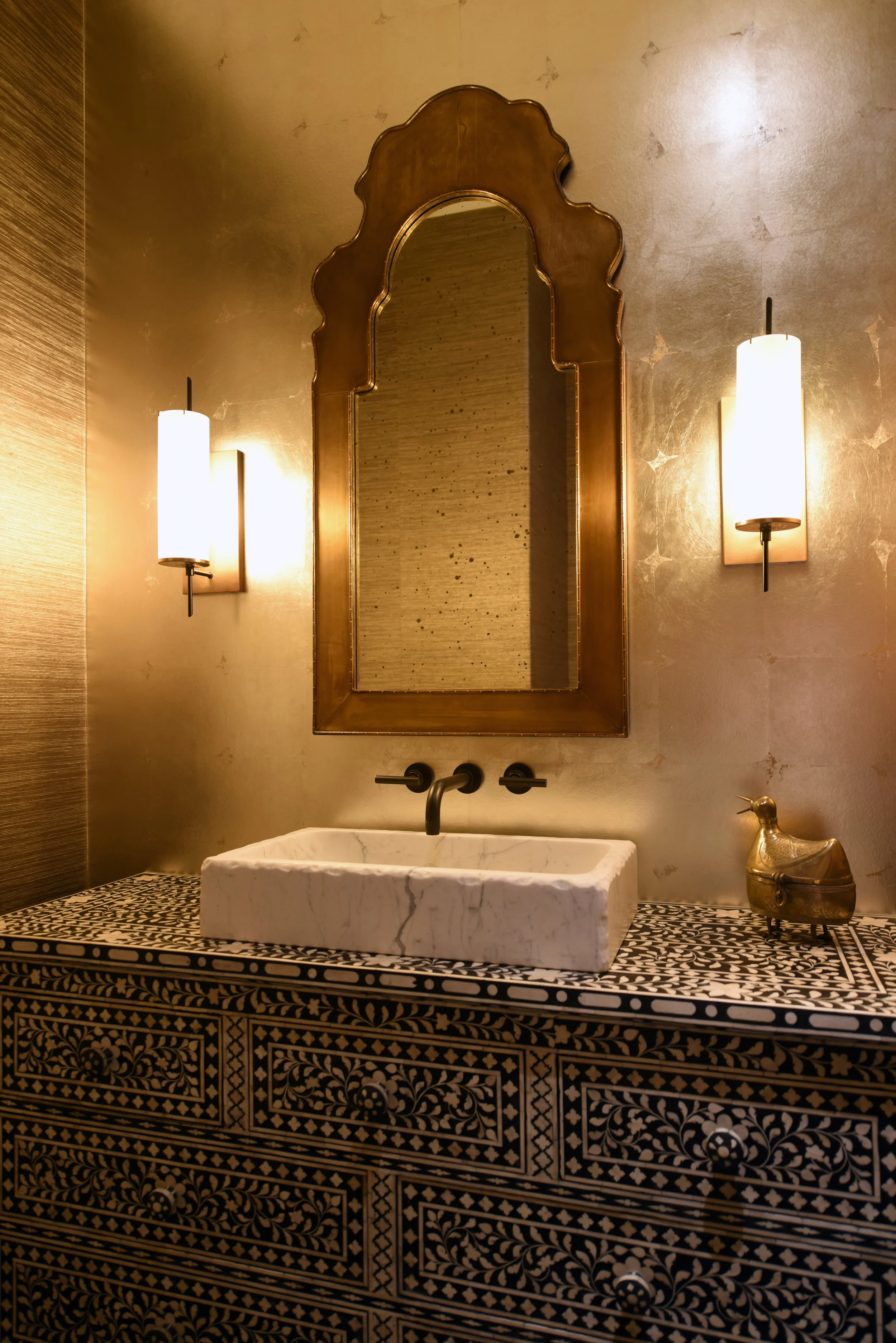 Bathroom vanity with a white stone sink, an ornate mirror, paired wall sconces, and a black-and-gold patterned counter.