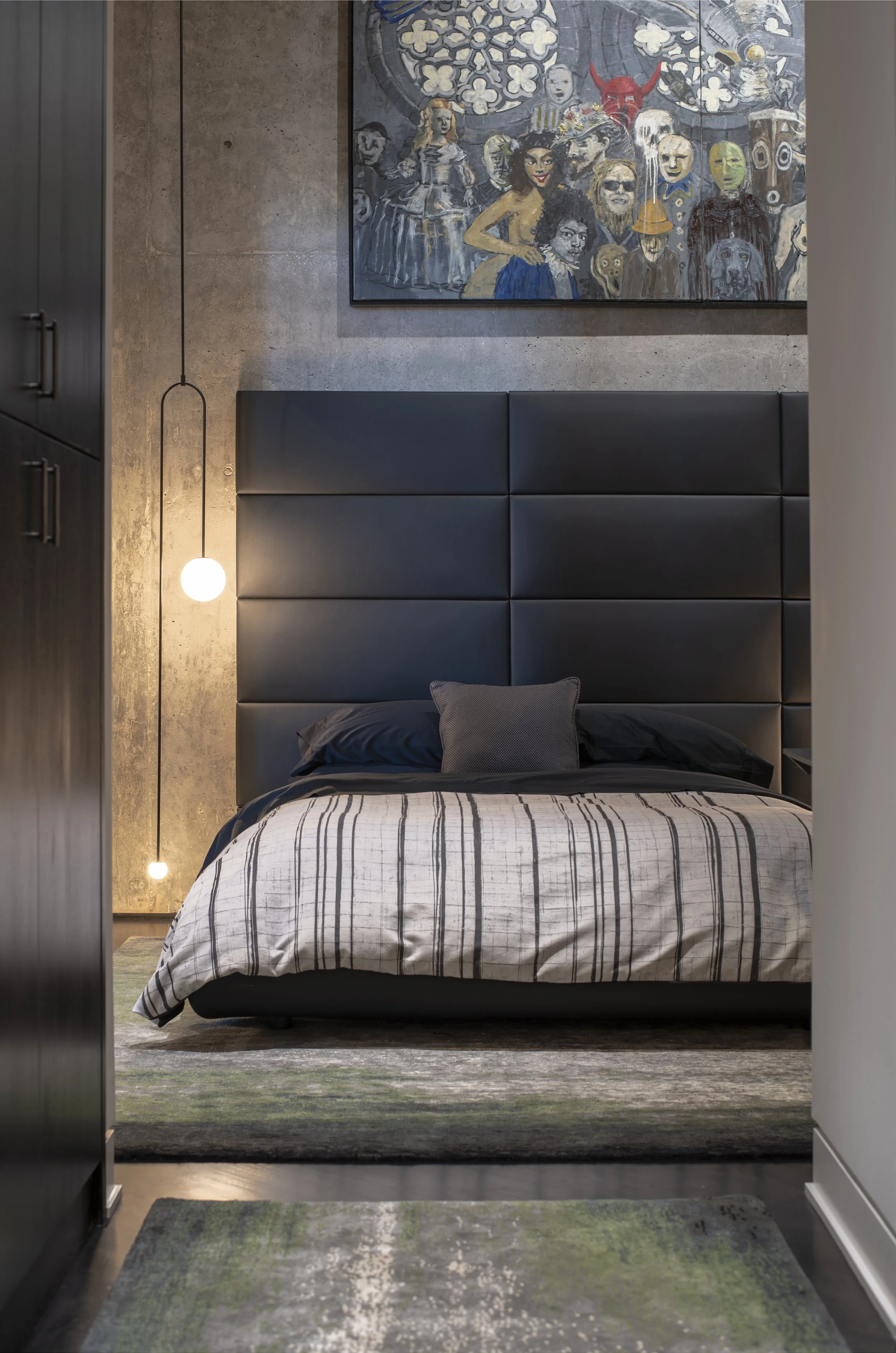 Bedroom with a black upholstered headboard, striped bedding, colorful pop-culture artwork on a concrete wall, and a green-and-gray rug.