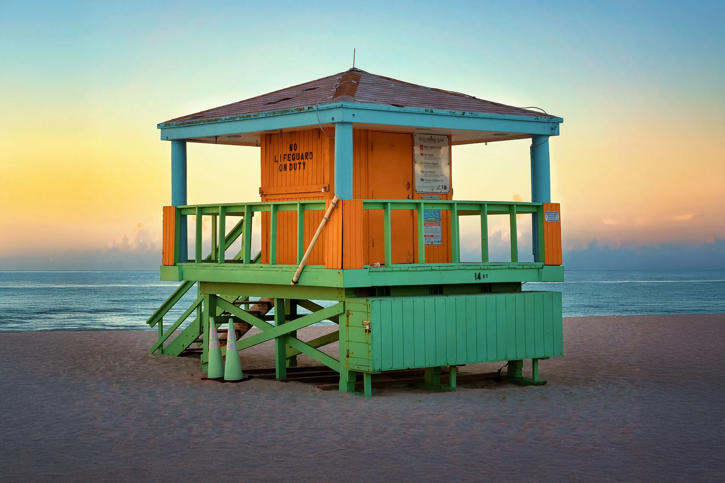 An orange and mint green lifeguard tower at 14th Street photographed straight on at dawn, with a soft pastel sky and calm ocean water behind it.