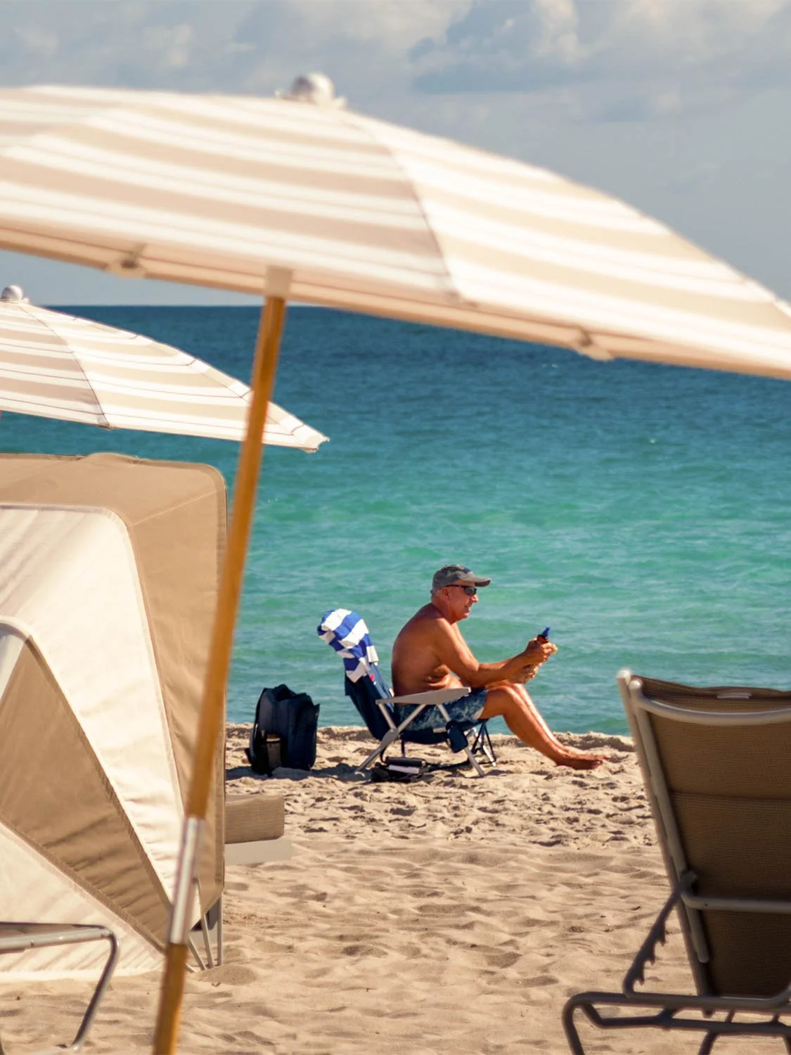 Man sitting in a beach chair at South Beach, shirtless with a cap, looking at his phone by turquoise water