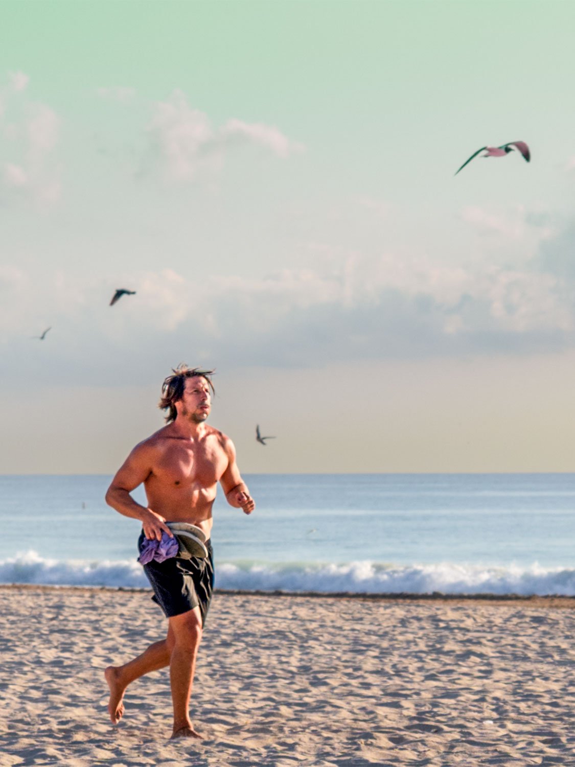 Shirtless man running barefoot on sandy beach with seagulls flying overhead and ocean waves in background