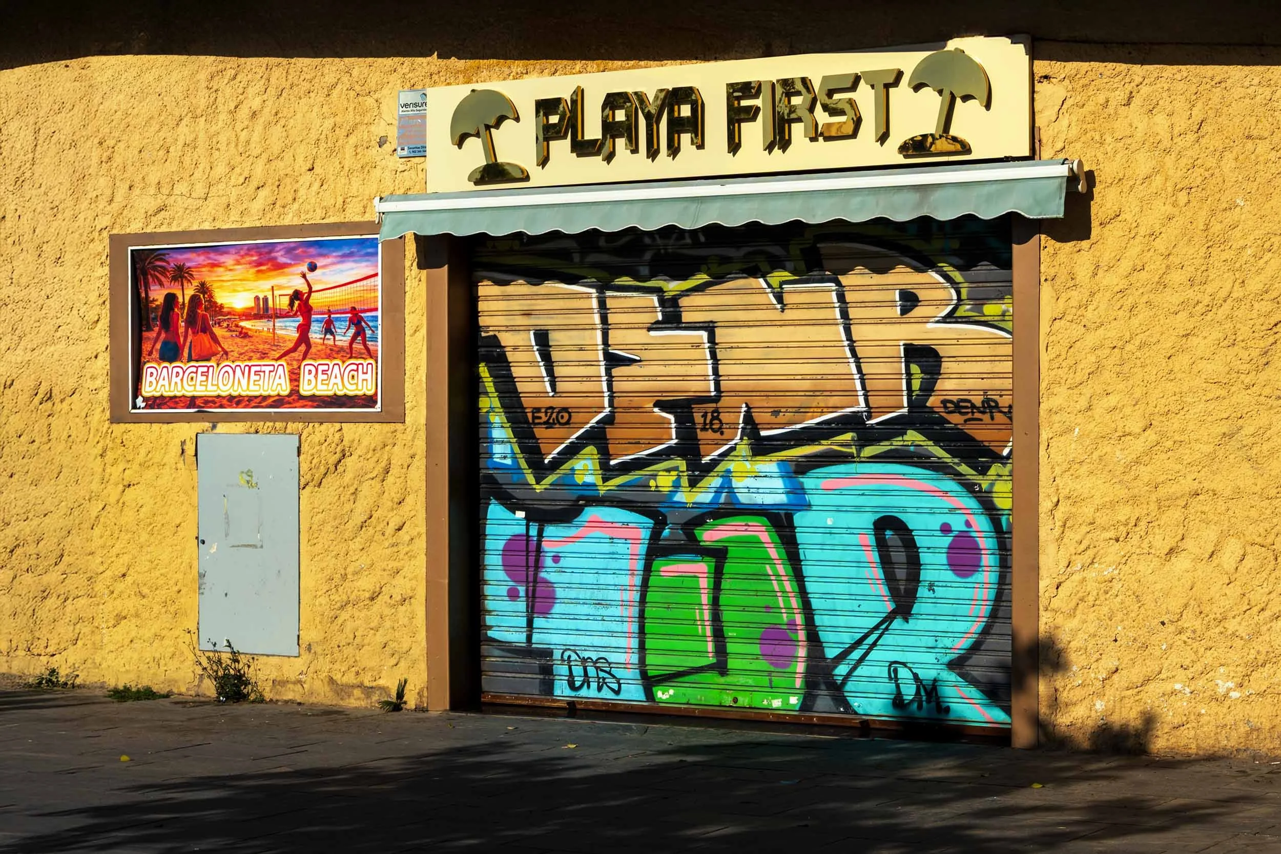 Closed Playa First beach shop with a graffiti-covered shutter and a Barceloneta Beach poster on a yellow stucco wall
