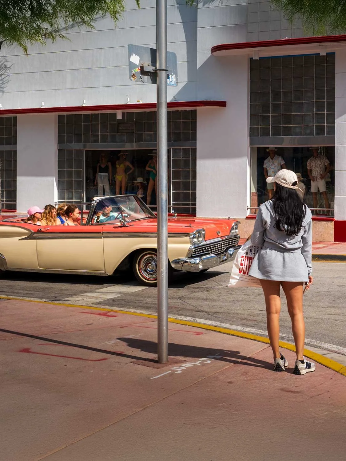 Woman in gray hoodie and cap watches a vintage red convertible cruise past an Art Deco building in Miami Beach