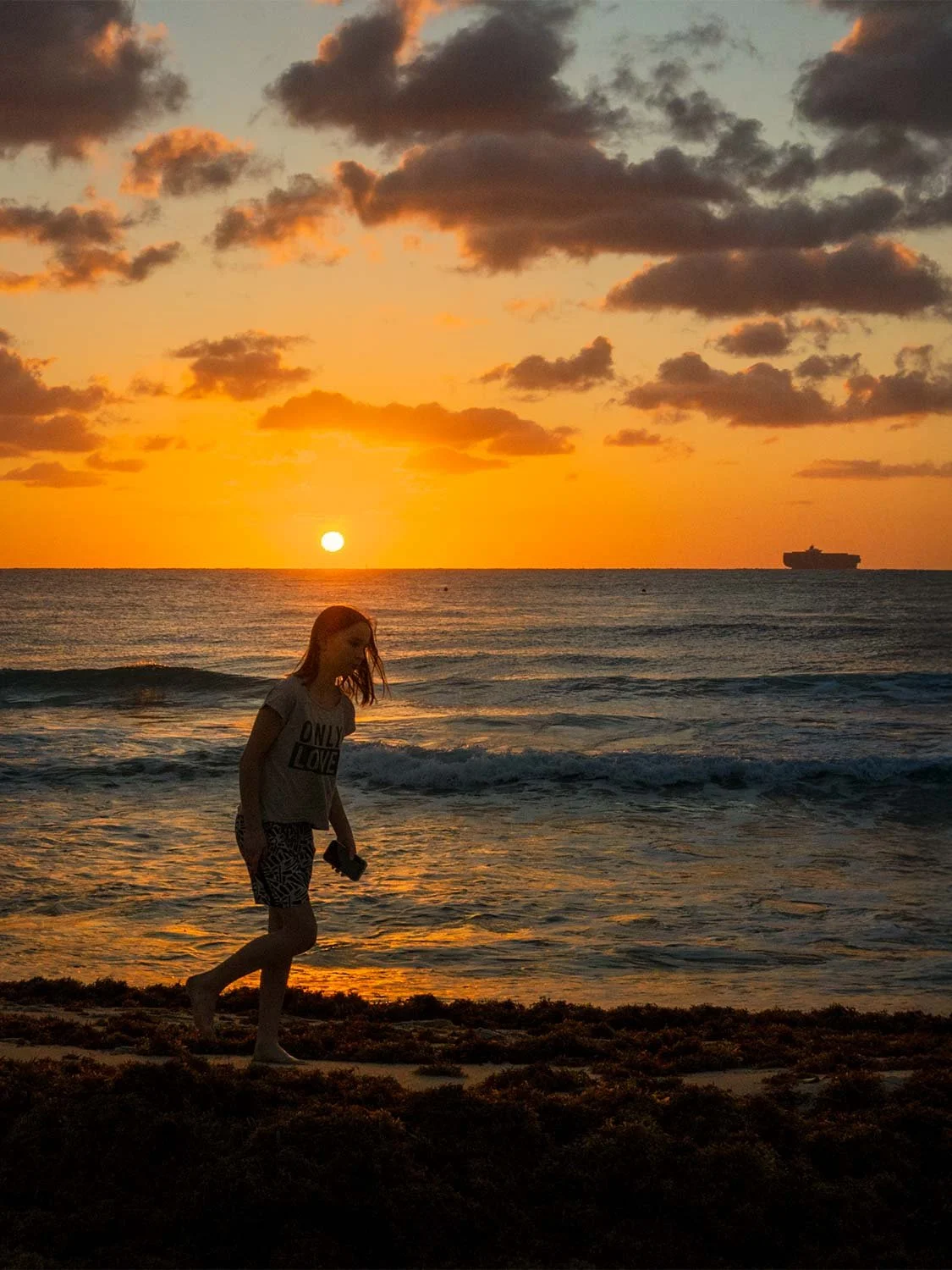 Woman walking barefoot along Sunrise South Beach at golden sunset with ocean waves and ship on horizon
