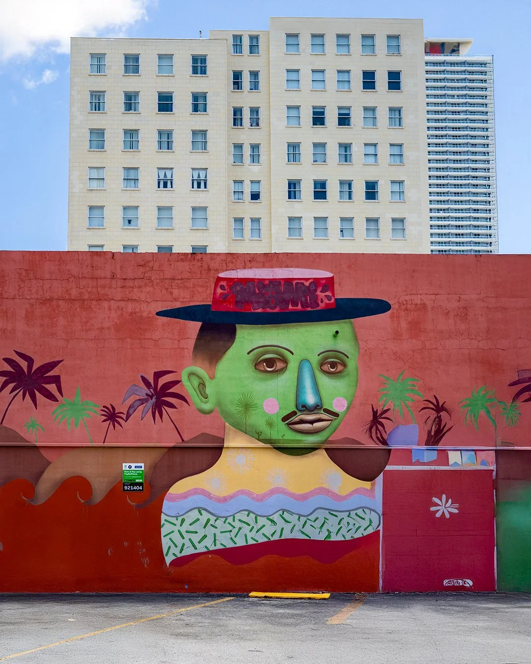 A green-faced figure in a boater hat on a red wall with palm trees, with an apartment block and a tower rising behind