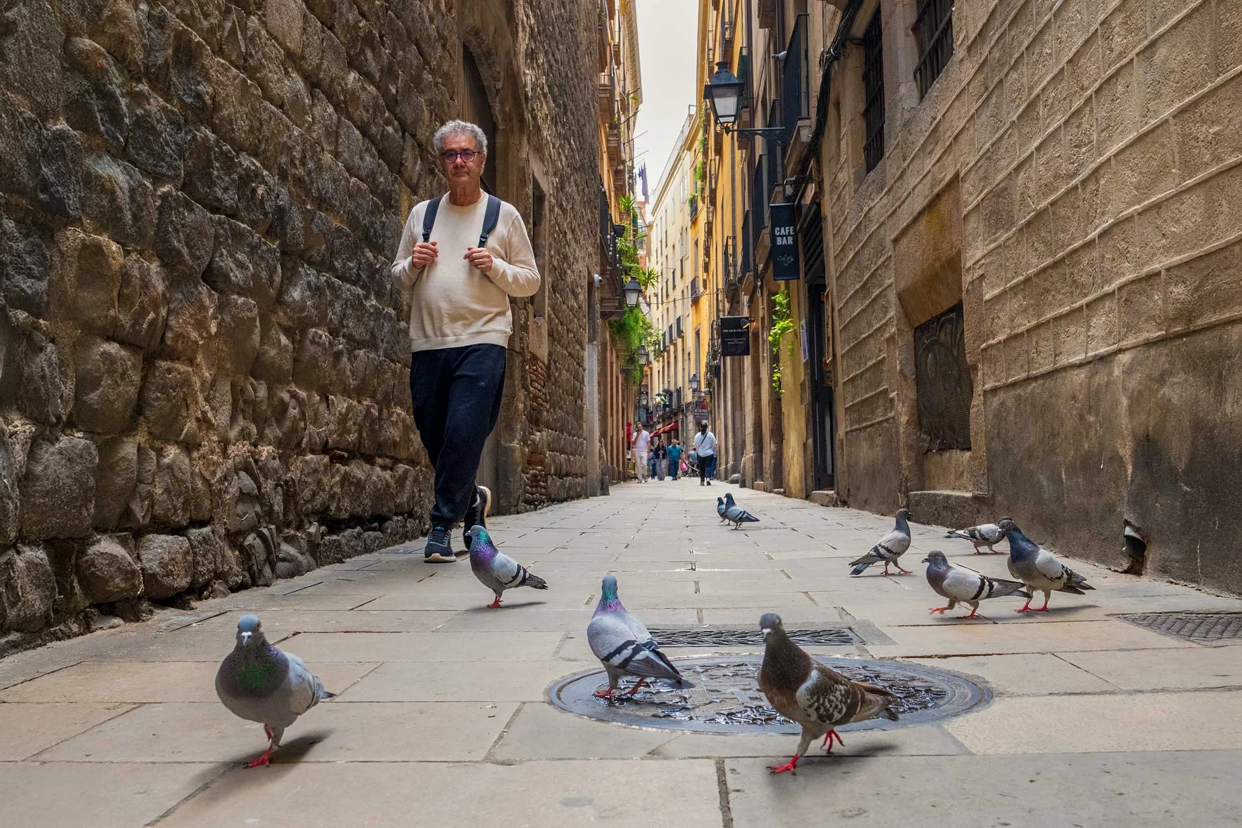 Man with a backpack walking down a narrow stone alley in Barcelona toward a flock of pigeons