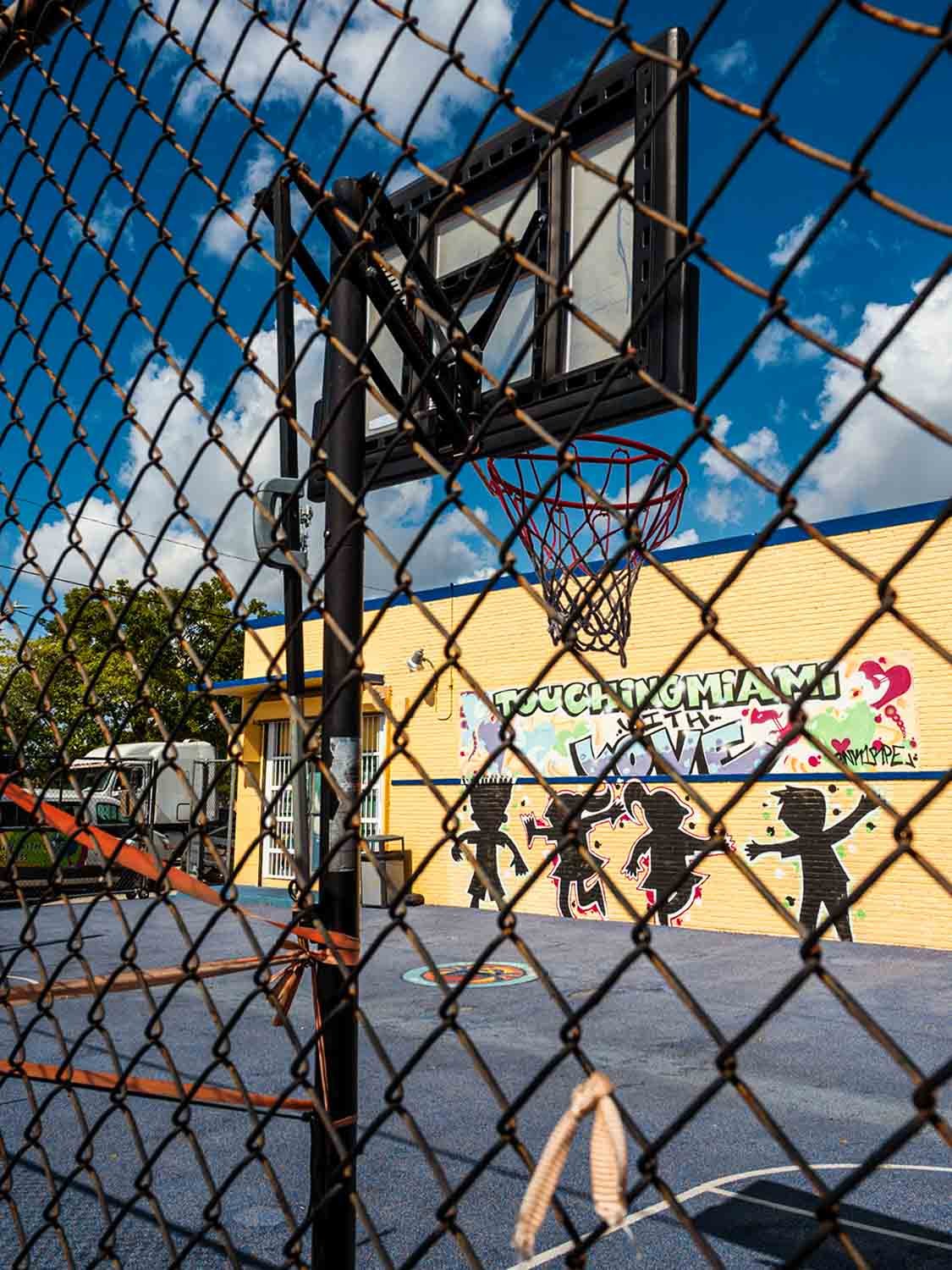 Downtown Miami basketball court viewed through a chain link fence, with the "Touching Miami with Love" mural on the wall behind