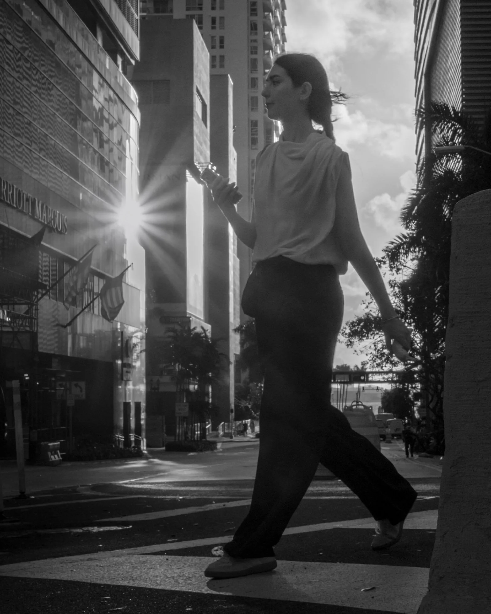 Black and white photo of a woman walking with her phone, sun flare at a building edge, Miami