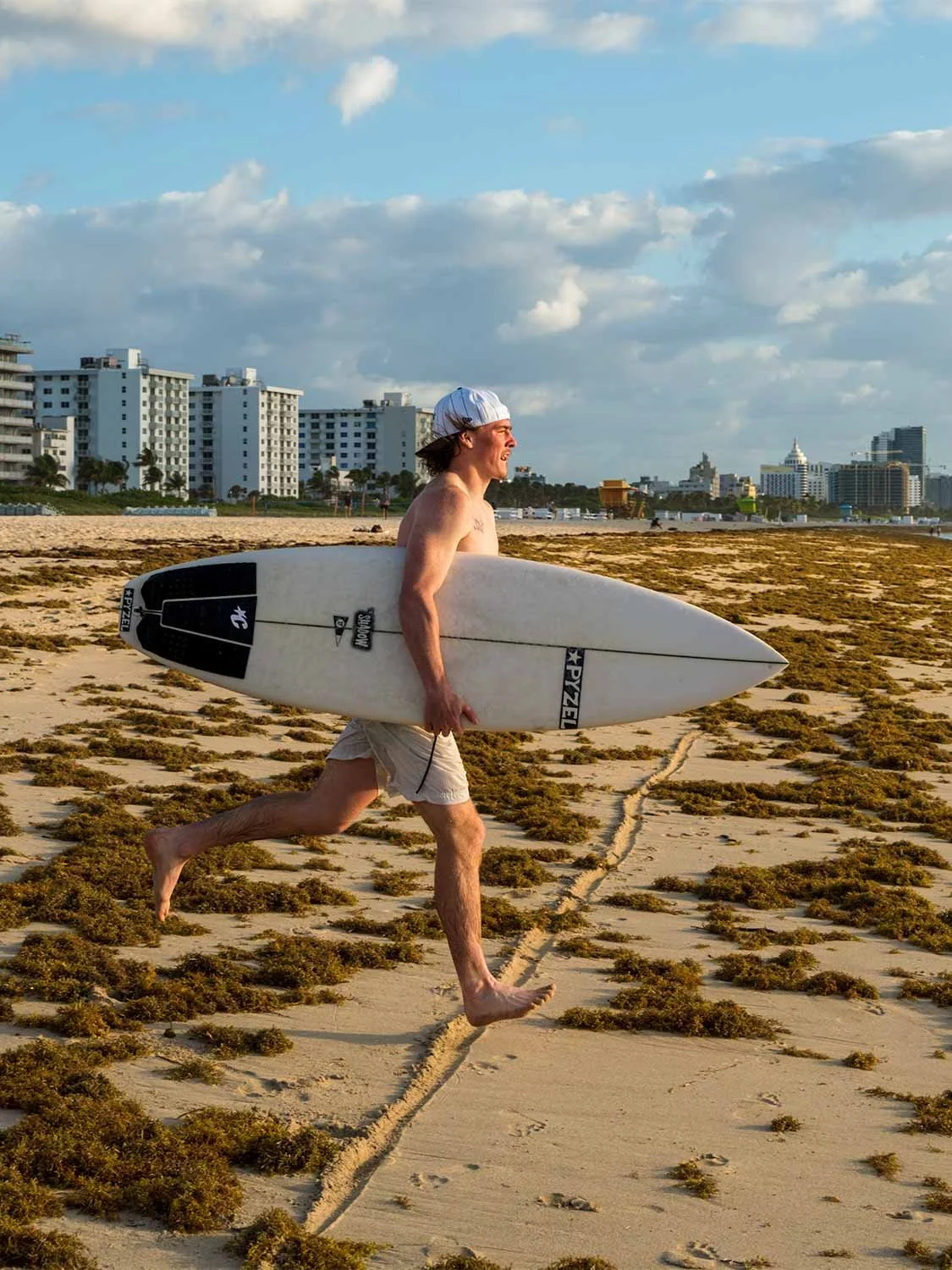 Shirtless surfer running barefoot on sandy Miami Beach carrying a white Pyzel shortboard under his arm