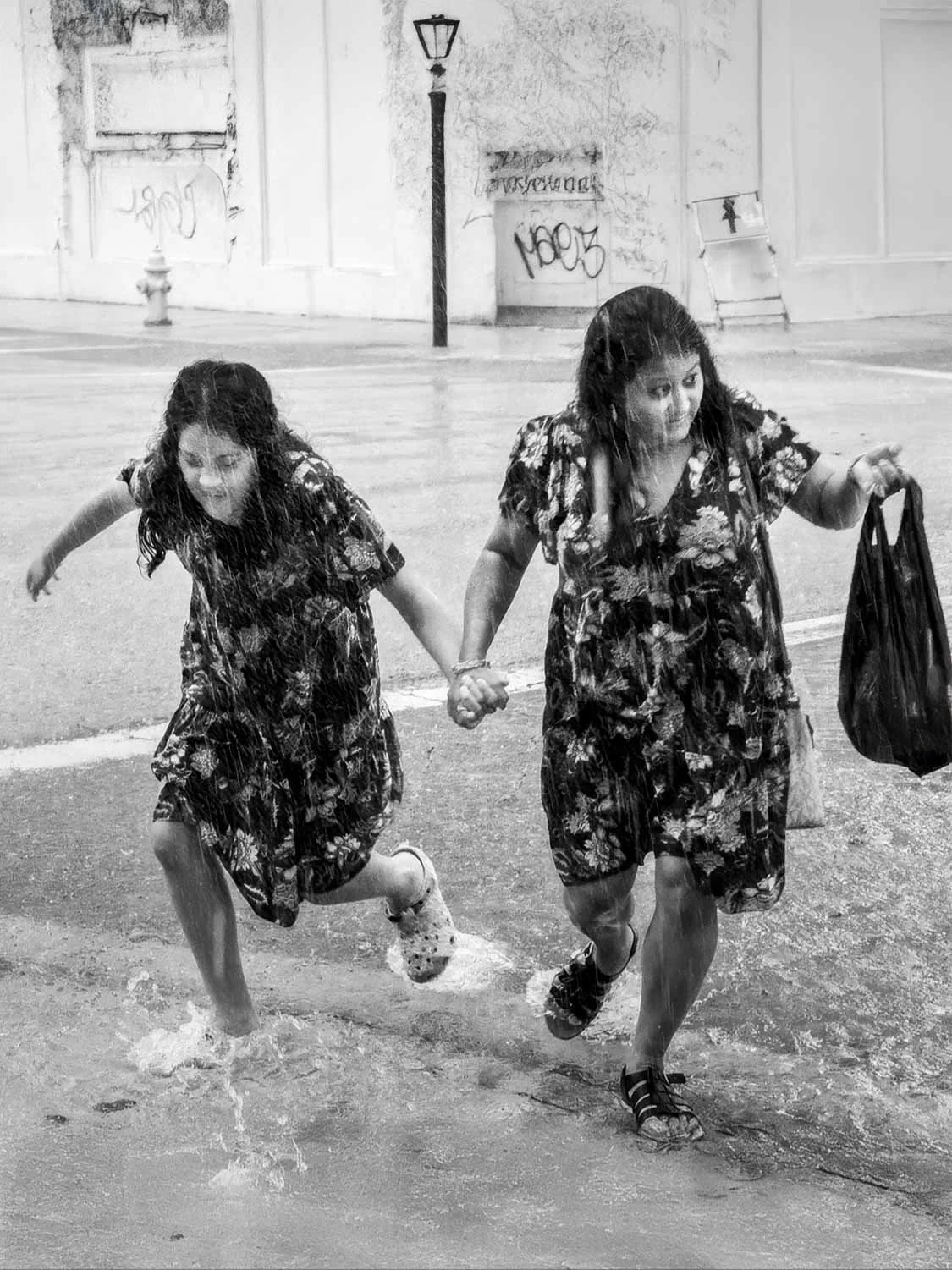Two women in matching floral dresses run hand-in-hand through heavy rain on a wet urban street