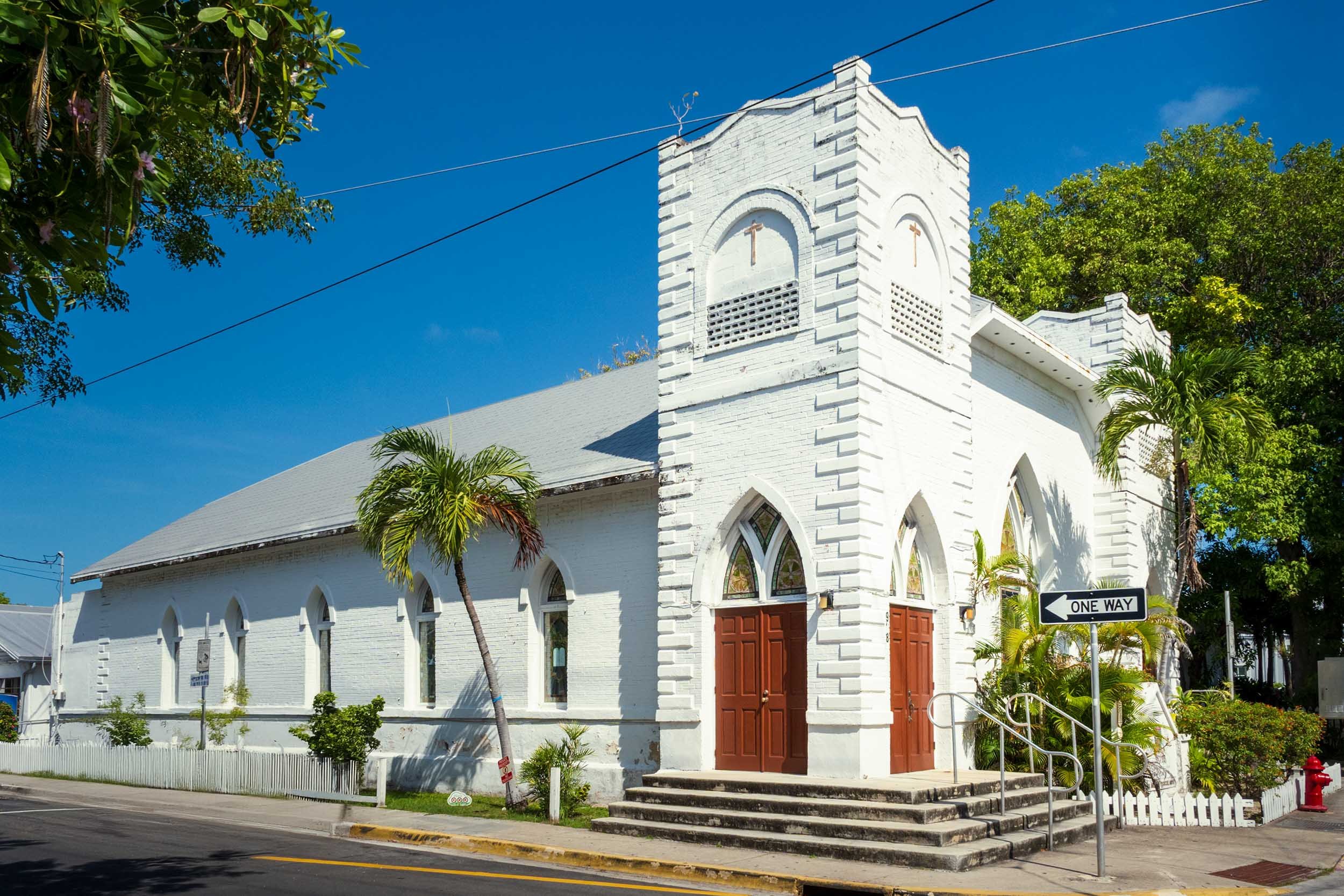 White historic church with Gothic arched windows and red doors surrounded by palm trees in Key West, Florida