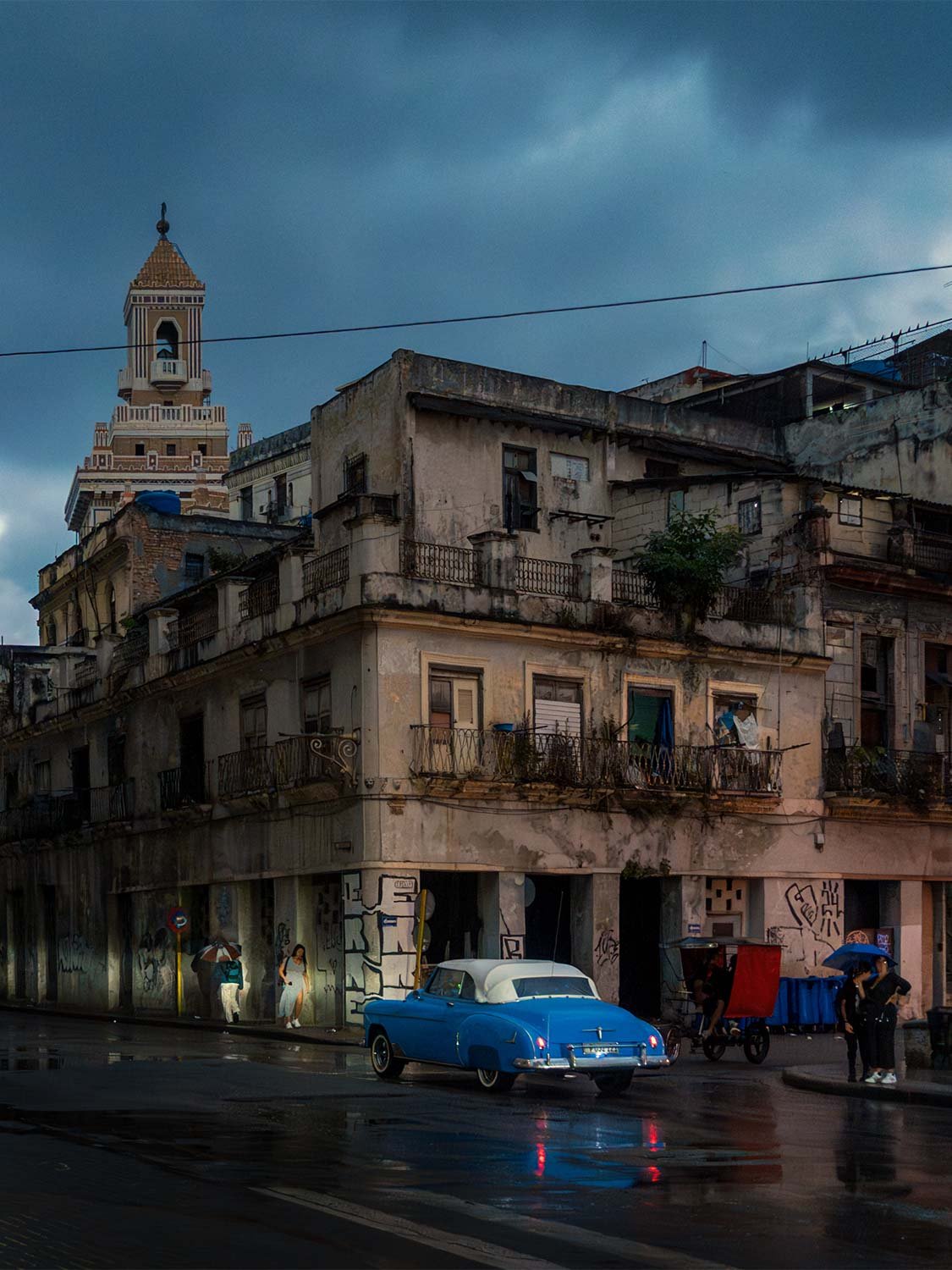 Blue classic convertible on a wet Havana street at dusk, the Bacardí Building tower rising behind weathered colonial buildings