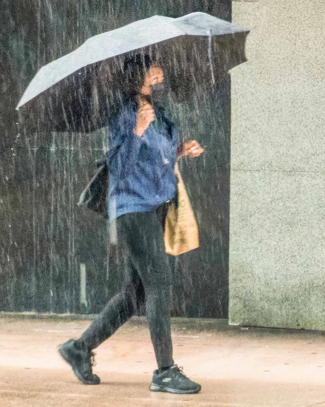 Person walking in heavy rain holding a light blue umbrella and yellow tote bag, wearing blue jacket and black pants