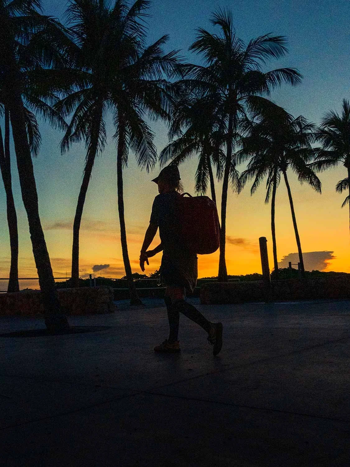 Silhouette of a man walking through Lummus Park at sunrise in South Beach, Miami, with warm light and palm-lined paths.