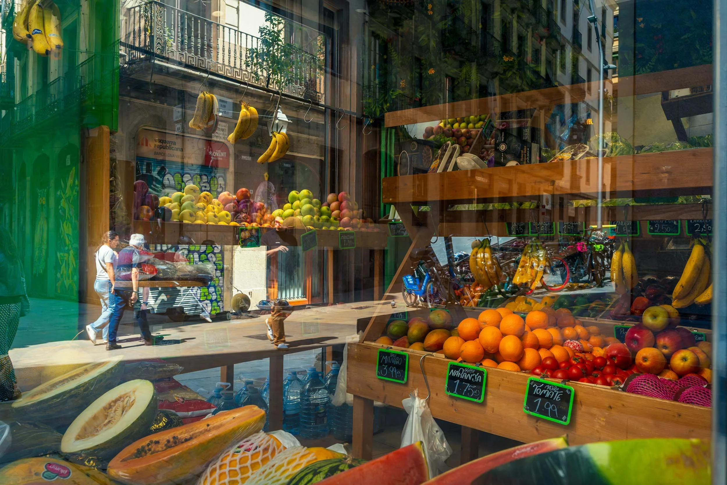 Oranges, melons and bananas in a fruit shop window, with pedestrians and old town balconies reflected in the glass