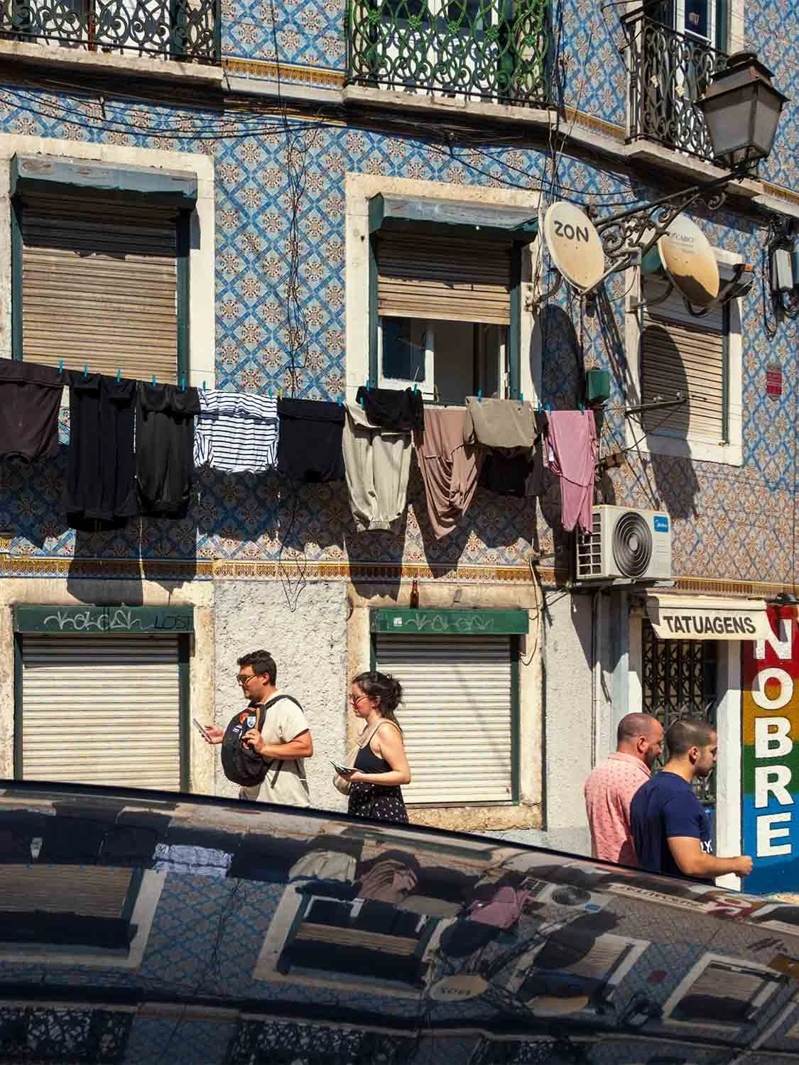 Laundry hanging across a blue tiled building in Lisbon as people walk past, the scene reflected in a car roof