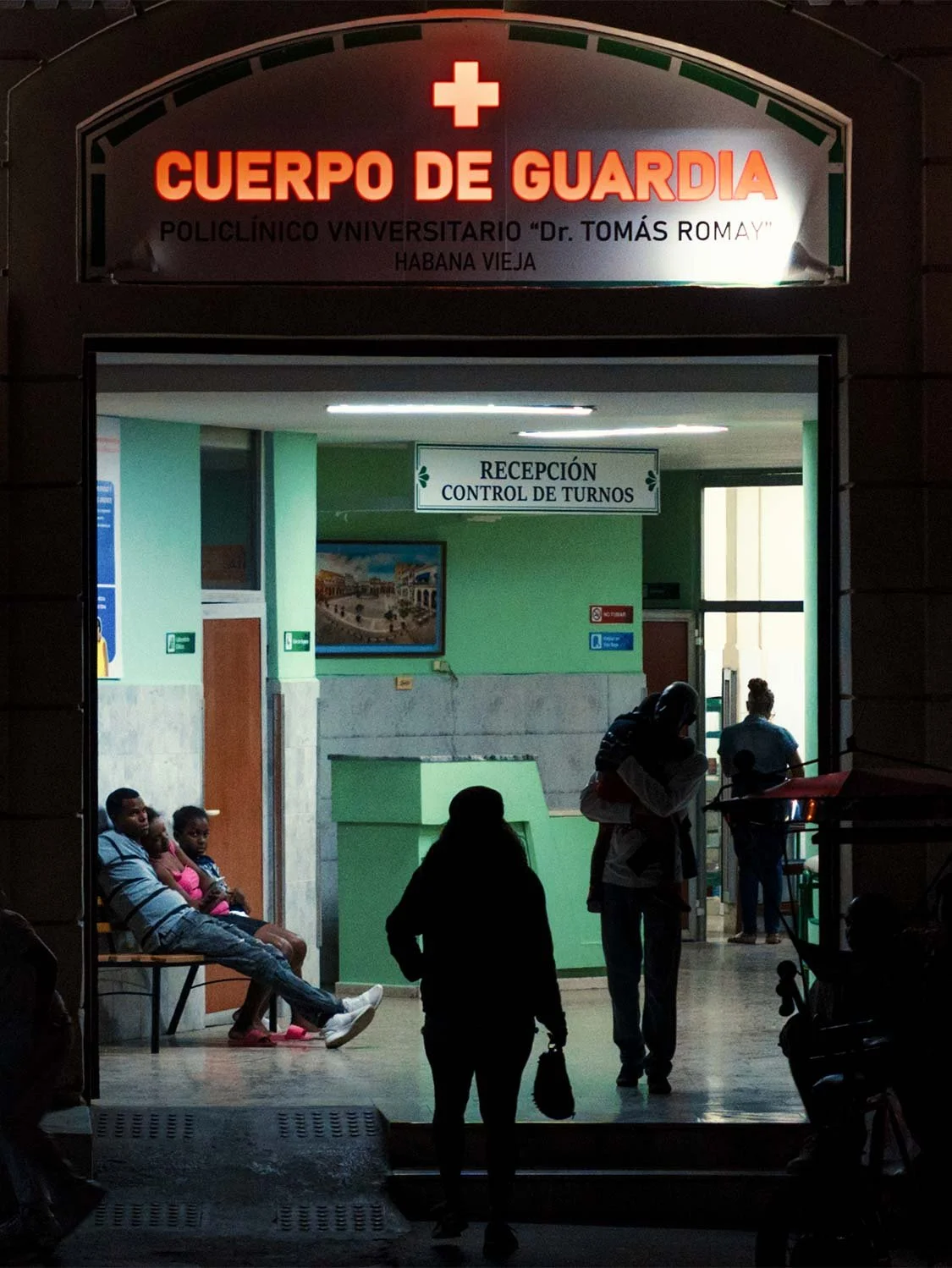 People waiting inside the lit entrance of the Cuerpo de Guardia emergency clinic in Old Havana at night