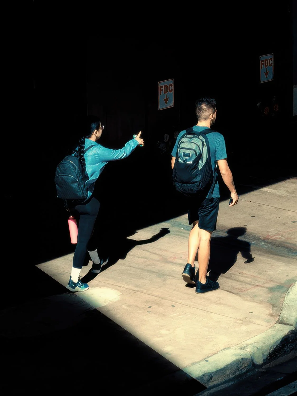 Two people with backpacks walking through a dramatic beam of sunlight on a shadowed downtown Miami sidewalk