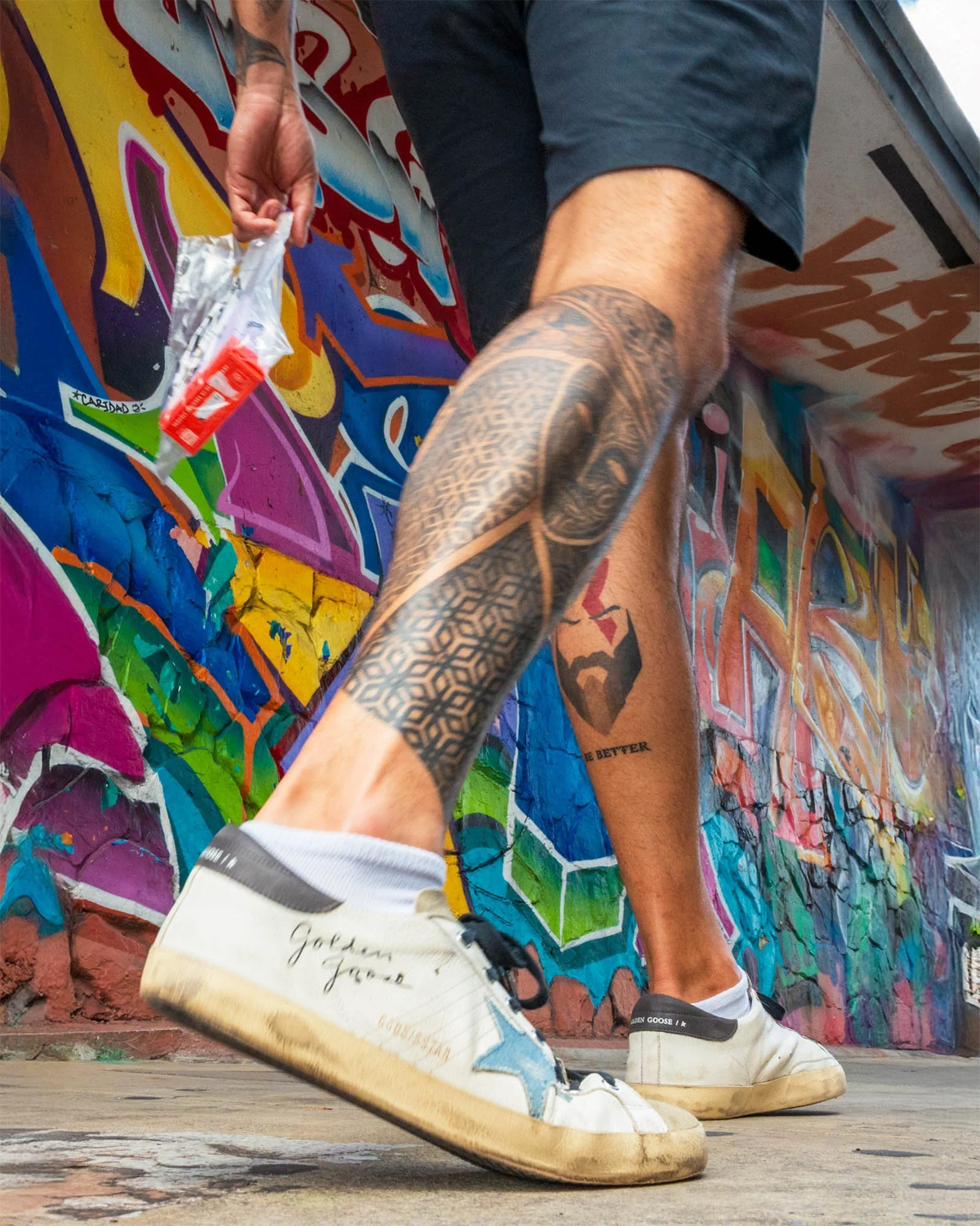 Worm's-eye view of a man with tattooed legs and worn white sneakers striding past a graffiti-covered wall in Miami.