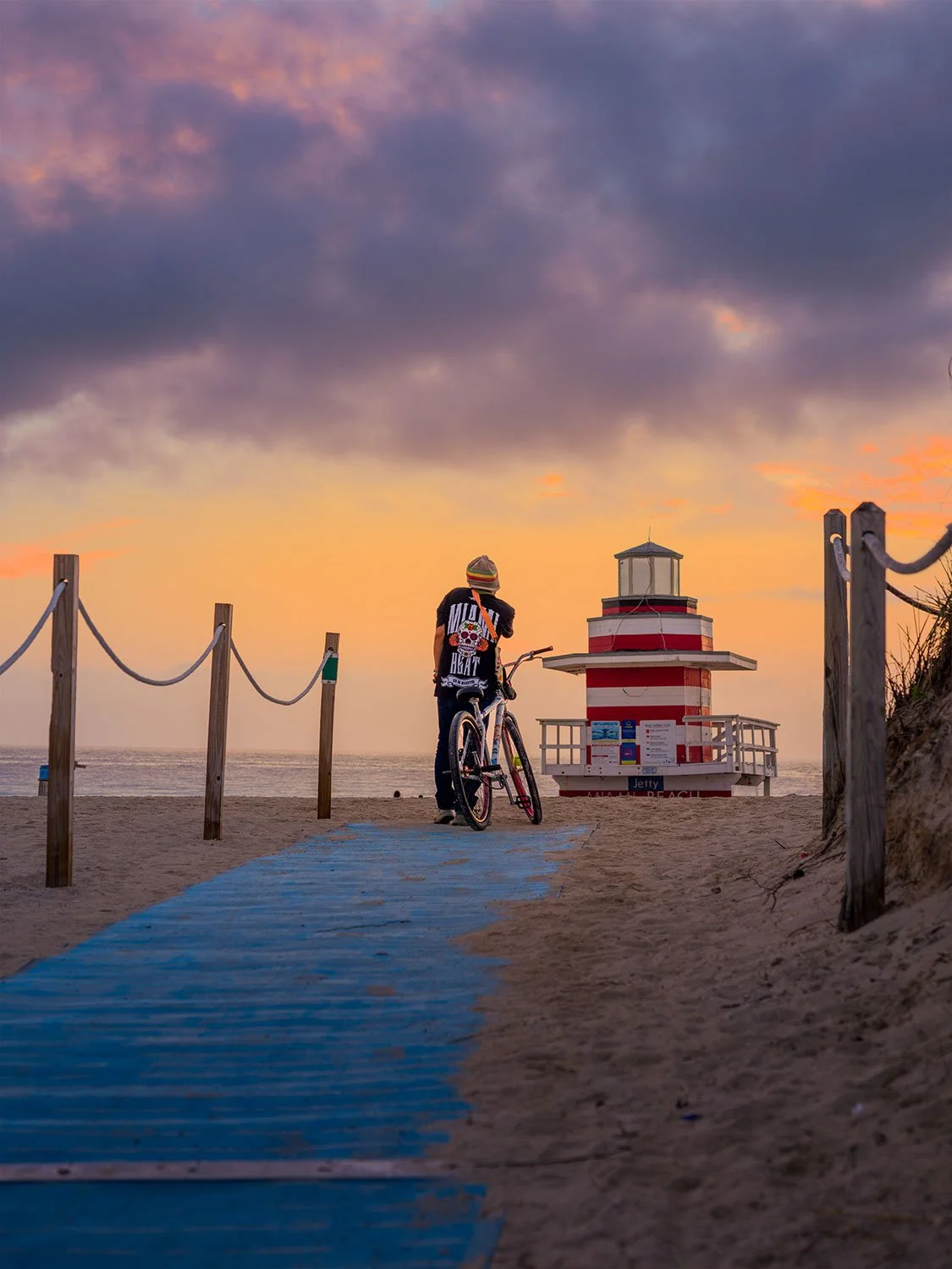 Man watching sunrise beside the Jetty Lifeguard Tower as soft golden light rises over the ocean and illuminates the shoreline.