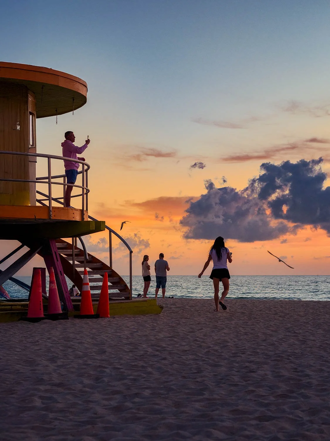 Silhouetted man standing on a lifeguard tower in South Beach Miami taking a photo of the sunrise over the ocean.