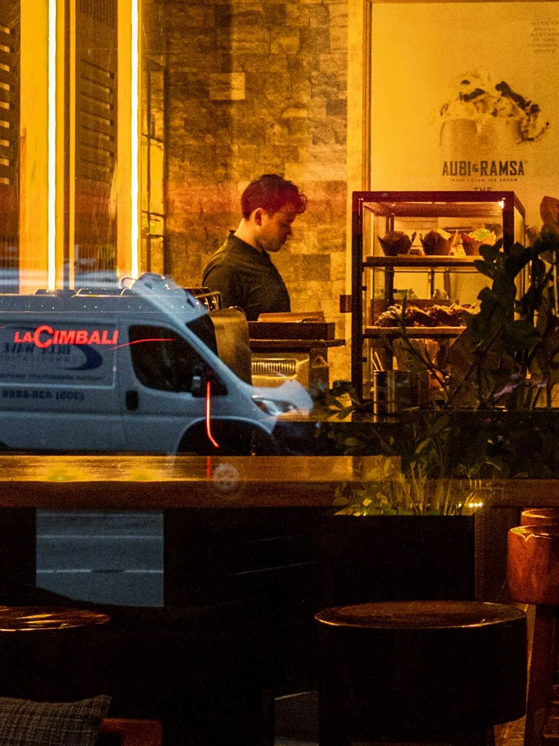 Restaurant worker inside a South Beach Miami café seen through a window with reflections of Ocean Drive at night and neon lights.