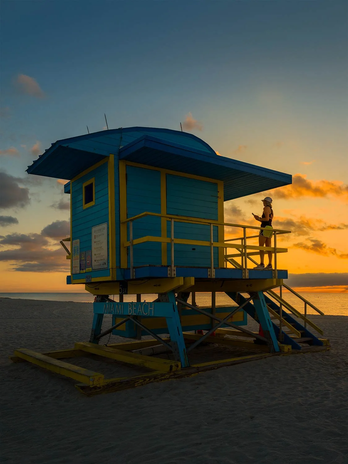 Person standing on a blue and yellow Miami Beach lifeguard stand at sunrise