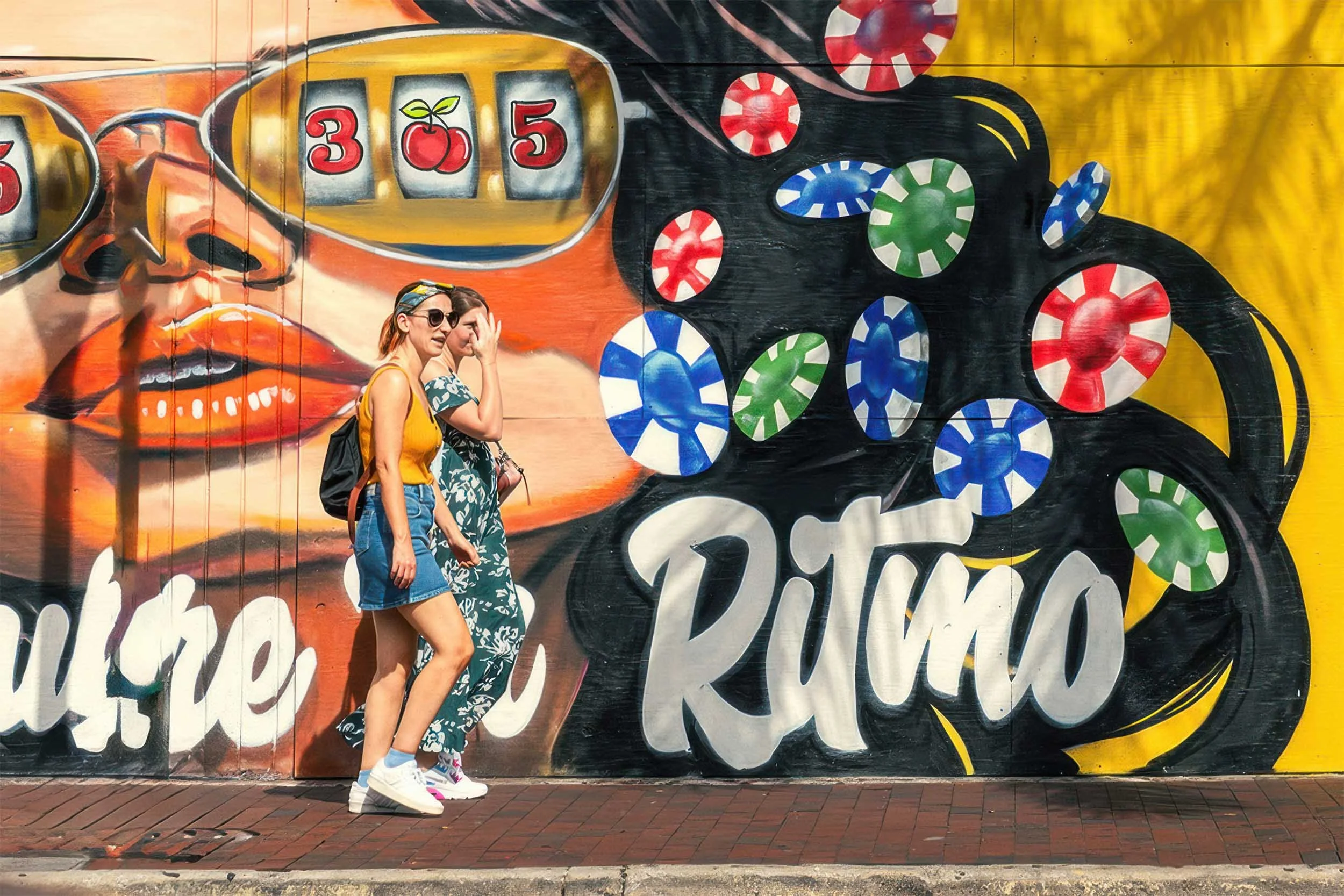 Two women walking past a casino-themed “Ritmo” mural with 305 slot-machine sunglasses in Miami