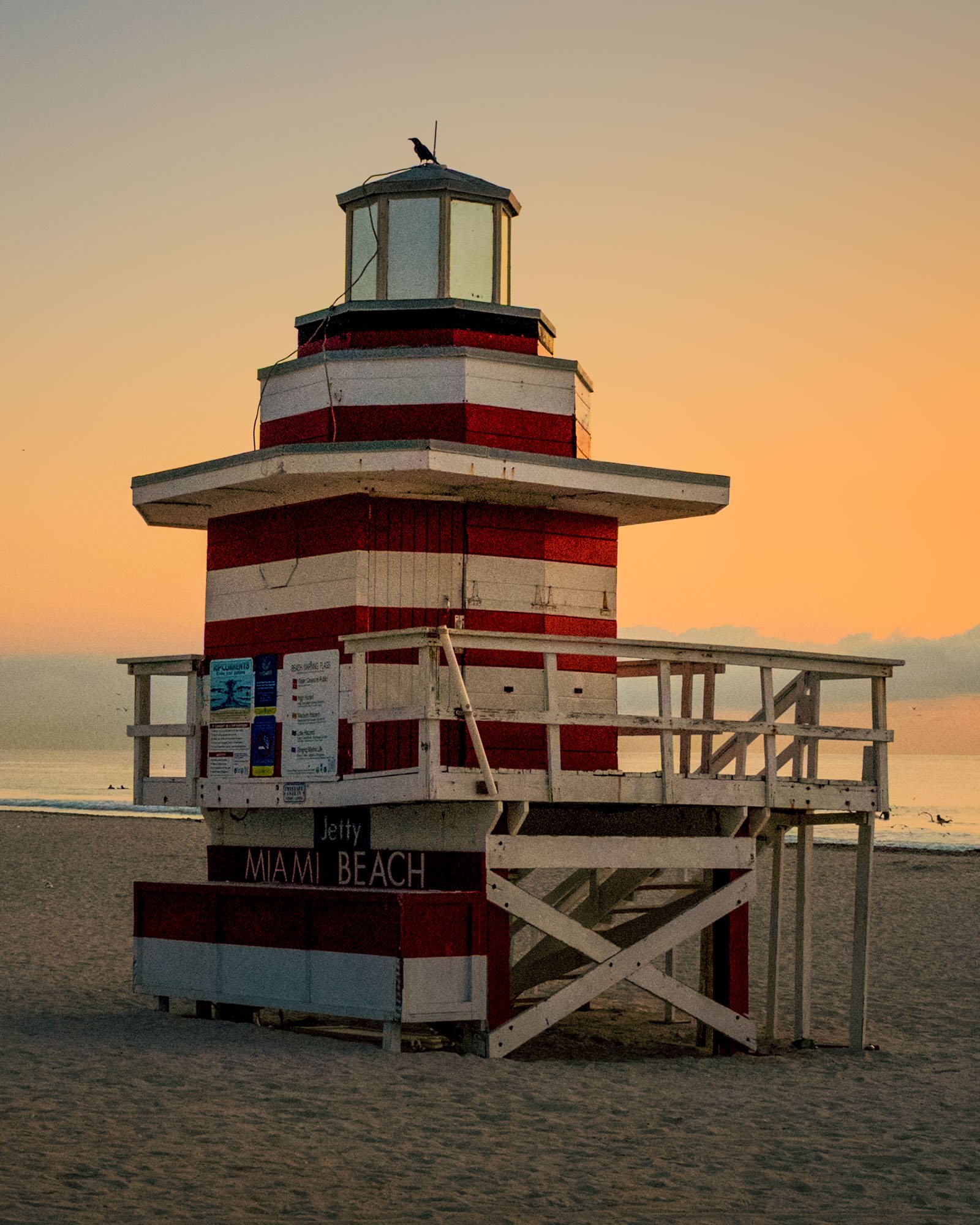 Weathered original Jetty lifeguard tower with a bird on the lantern and a Miami Beach sign on the deck, golden sky over the ocean