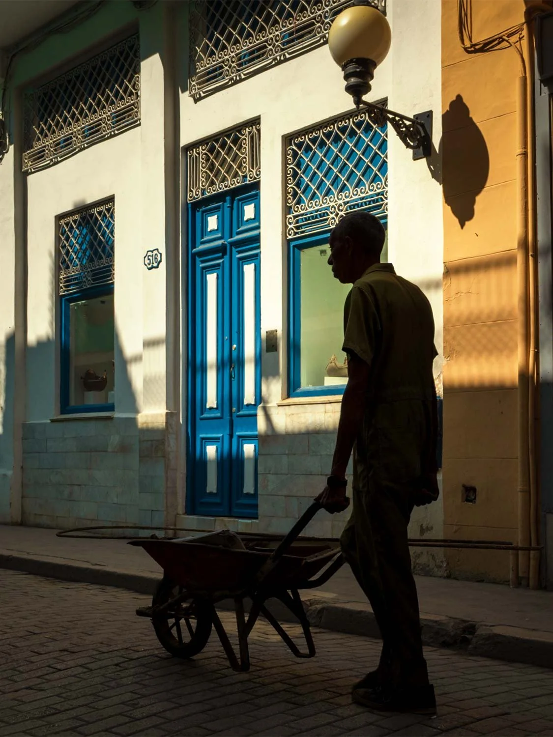 Silhouette of worker with wheelbarrow on cobblestone street, colonial architecture, blue doors, Havana Cuba street photography.