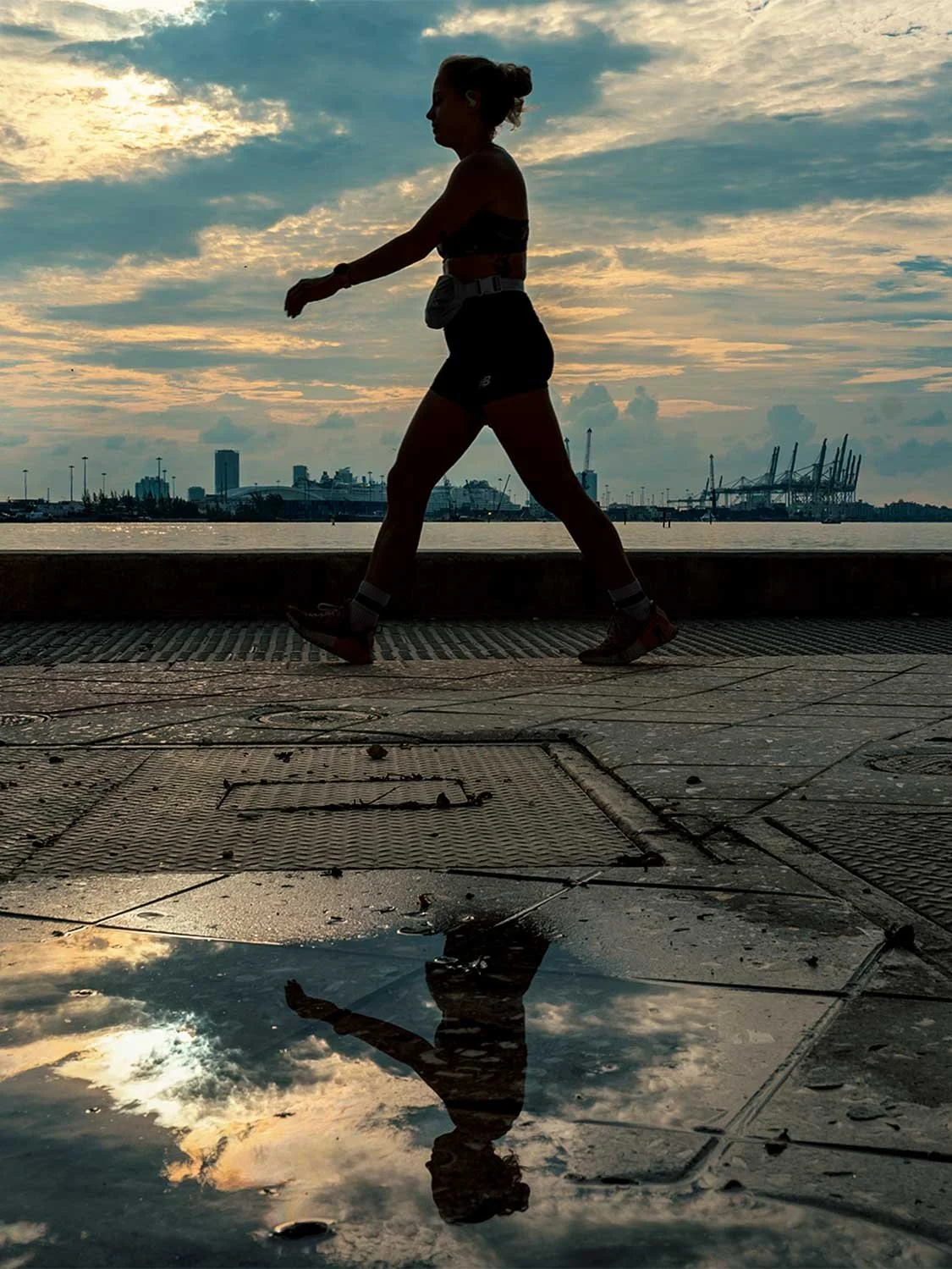 Female athlete walking with ankle weights at sunset waterfront, city skyline background, puddle reflection fitness photo.