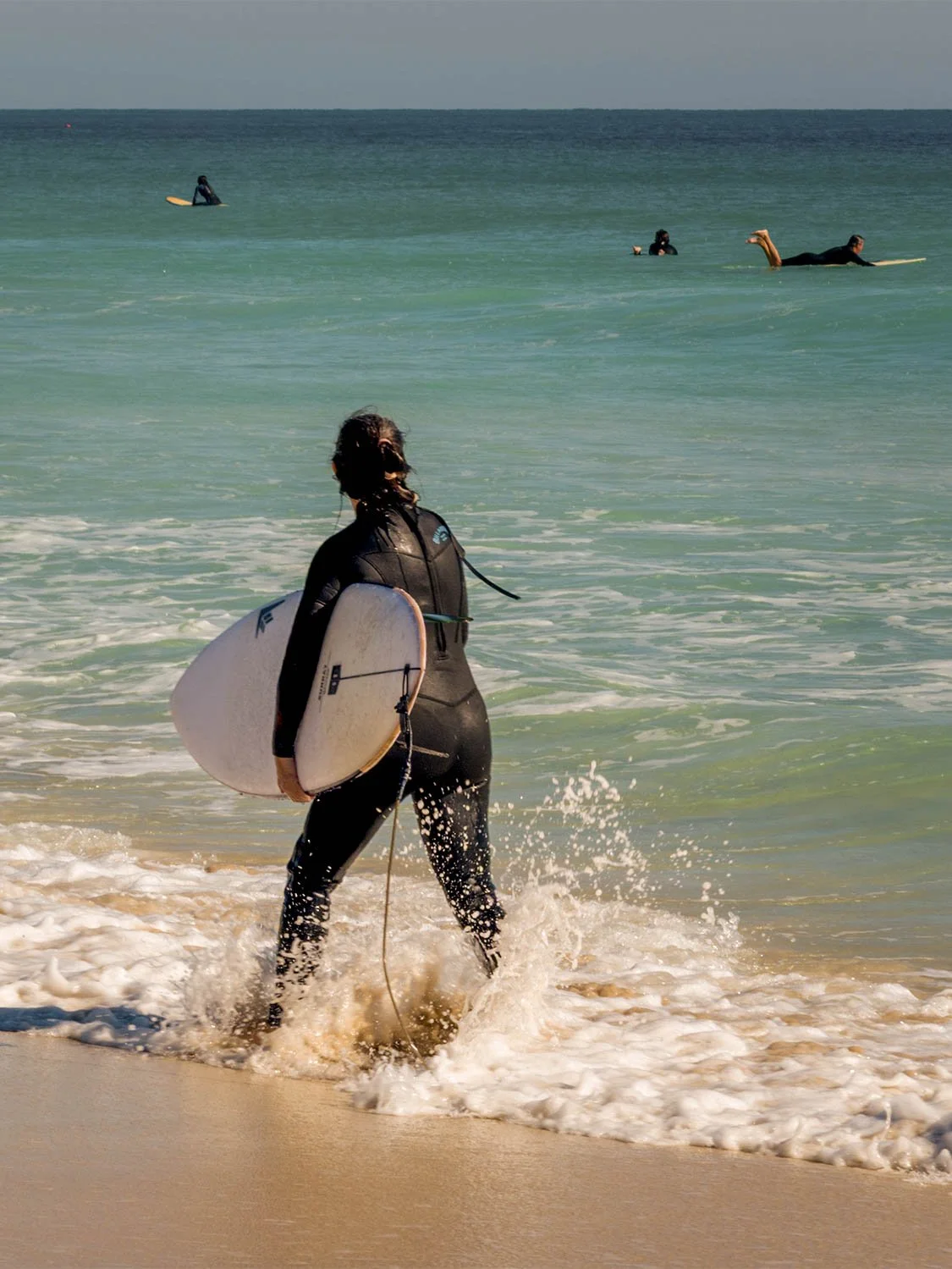 Surfer in black wetsuit carrying white surfboard wading into turquoise ocean waves on a sandy beach