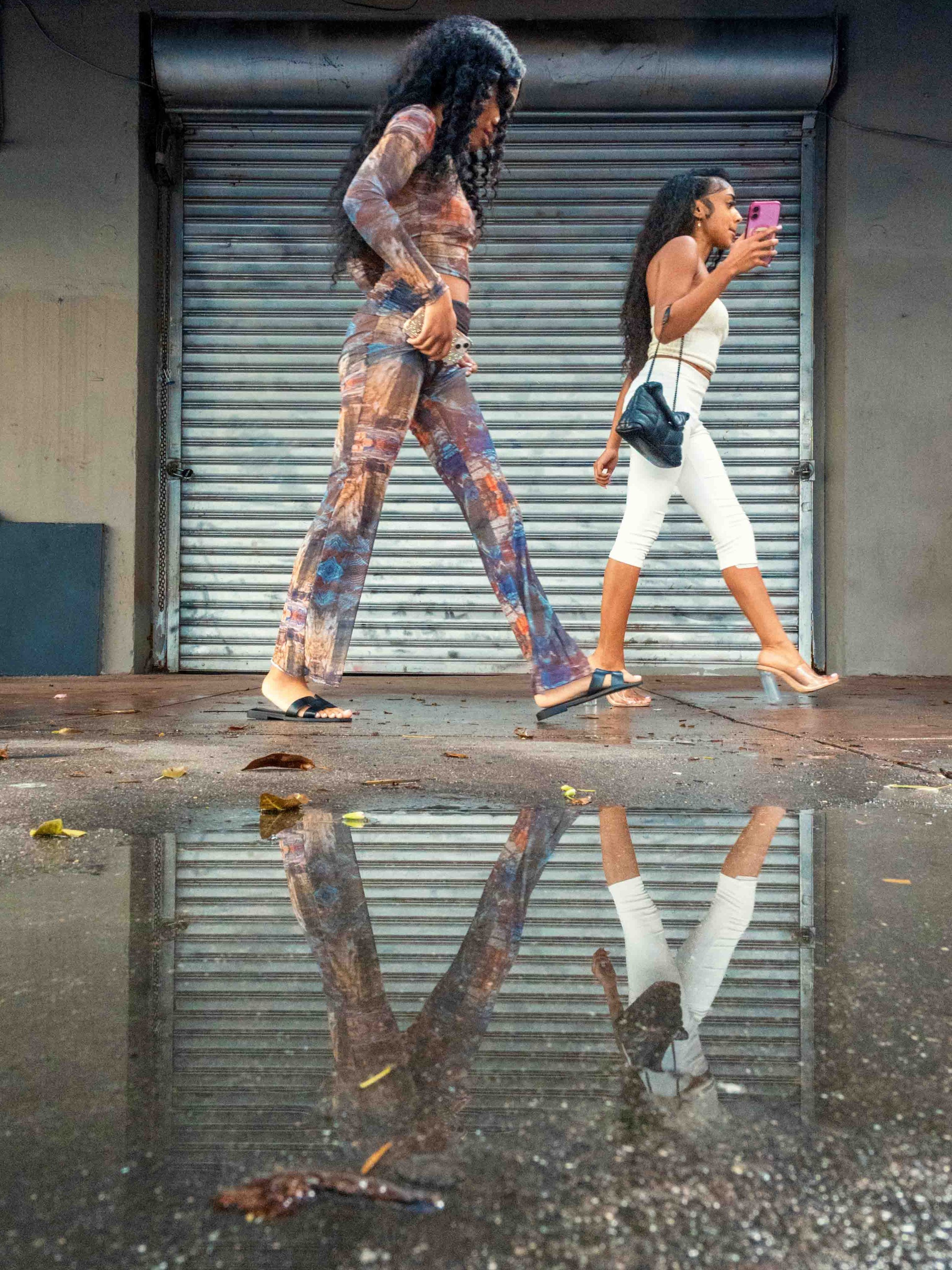 Two women walking past a metal roll-up door on a wet Downtown Miami sidewalk, their reflections caught upside down in a puddle from a camera set on the ground.