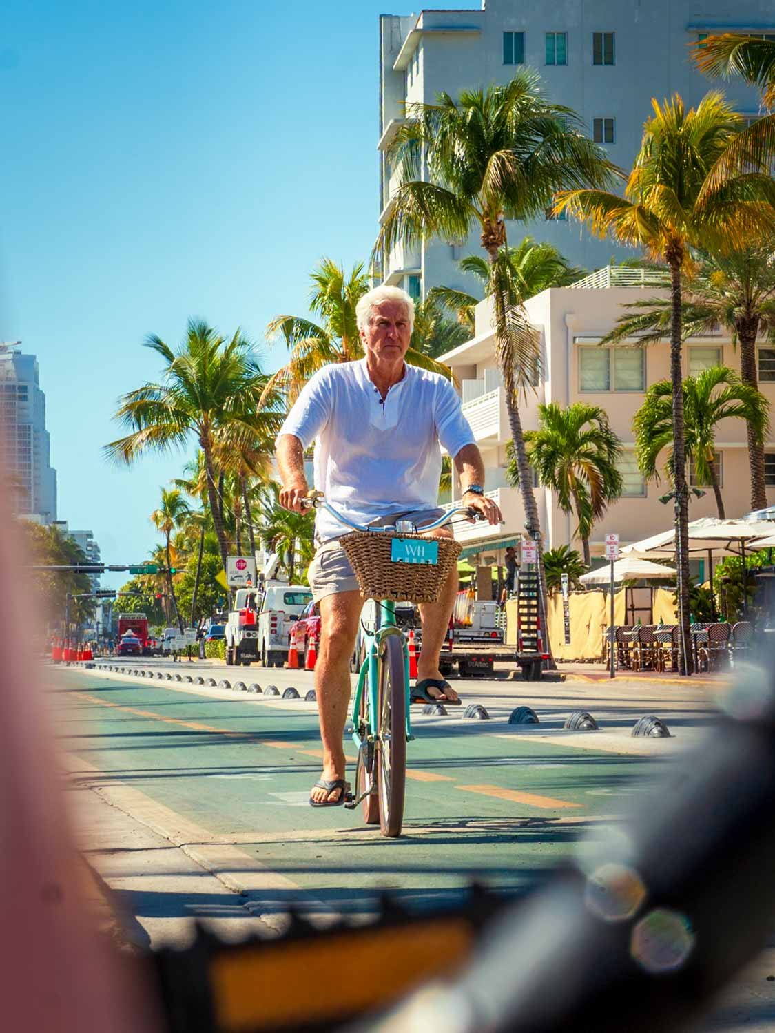 Senior man with white hair rides a teal cruiser bicycle with wicker basket on a sunny Miami Beach boulevard